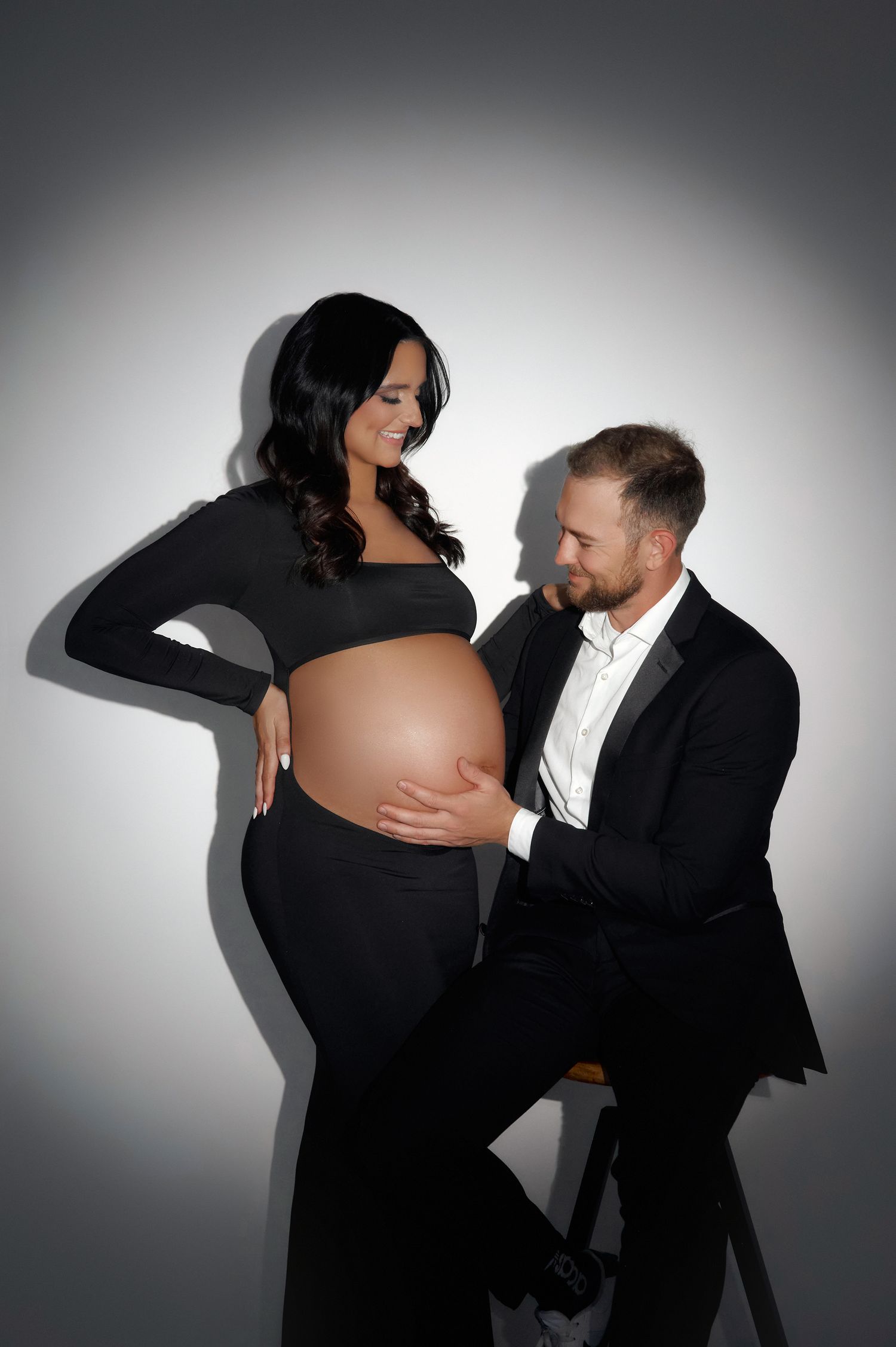 Pregnant woman in black outfit with partner in suit tenderly touching baby bump in studio portrait.
