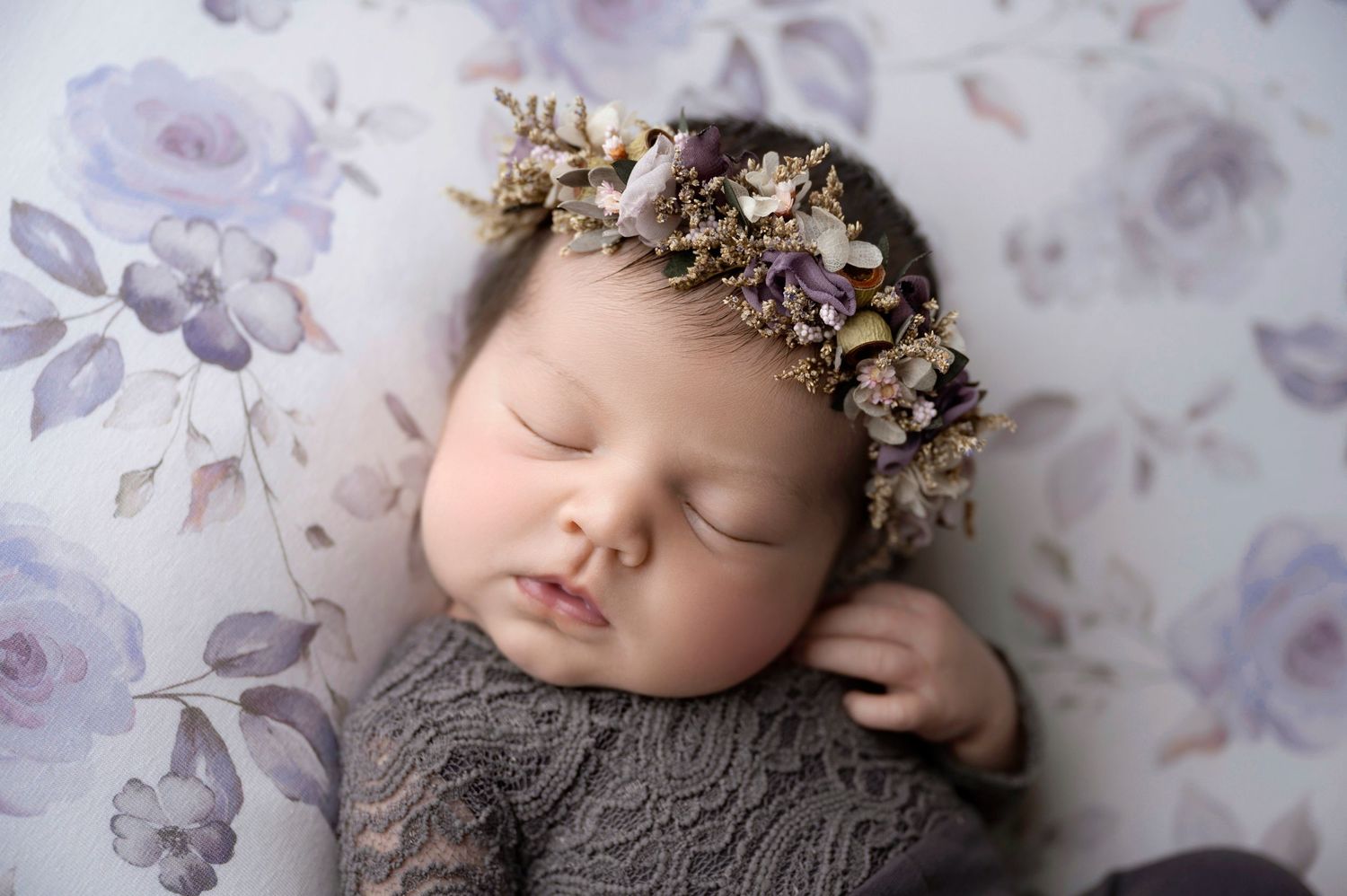 Sleeping newborn baby wearing a purple floral crown, dressed in gray lace, on a soft floral background.