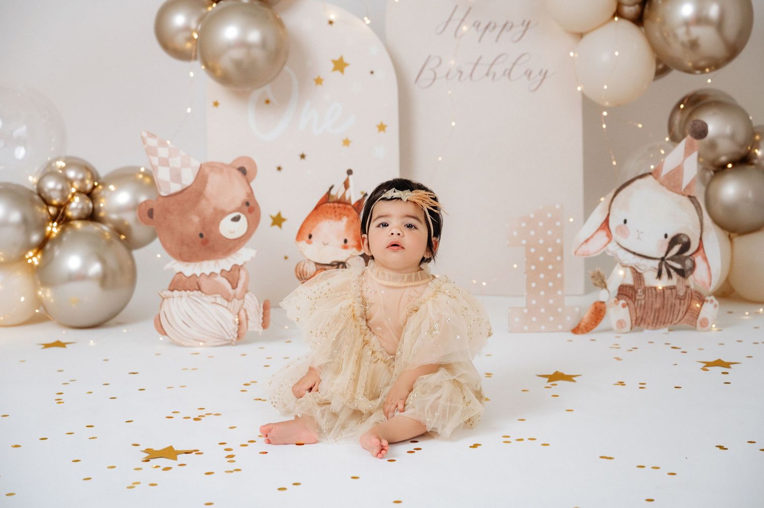 Baby girl in cream dress sits before Happy Birthday backdrop with gold balloons and animal cutouts at first birthday party.