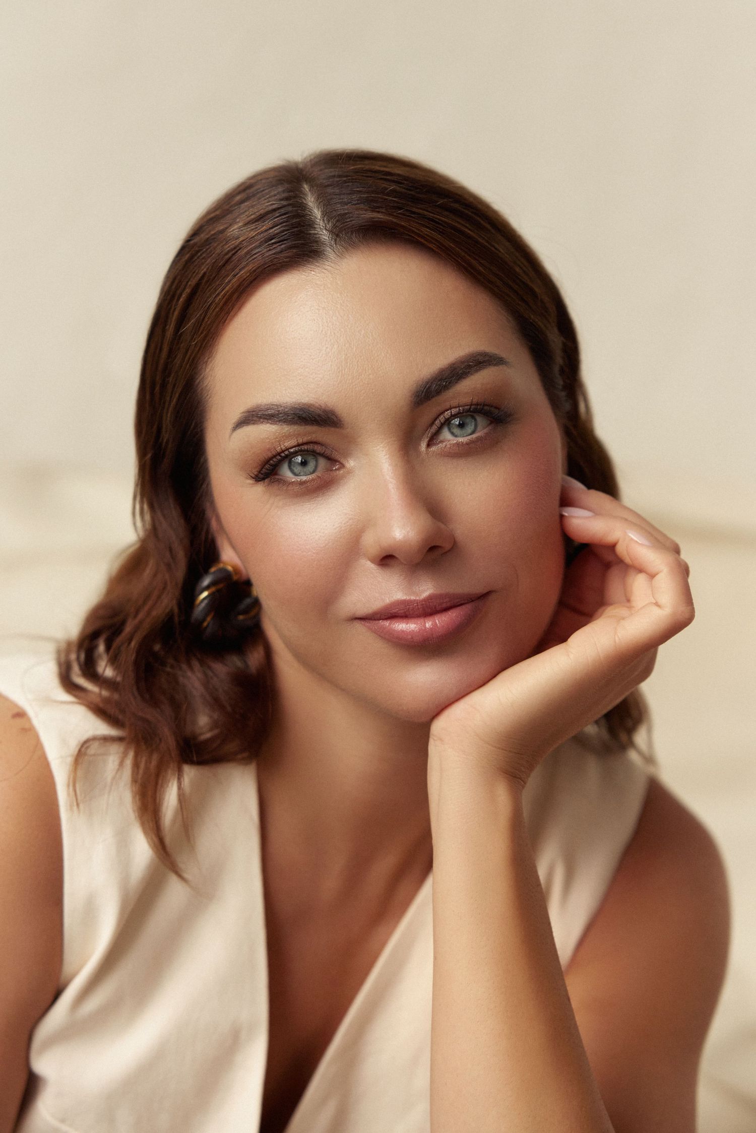 A brunette woman with blue eyes and elegant makeup rests her chin on her hand, wearing a cream sleeveless top.