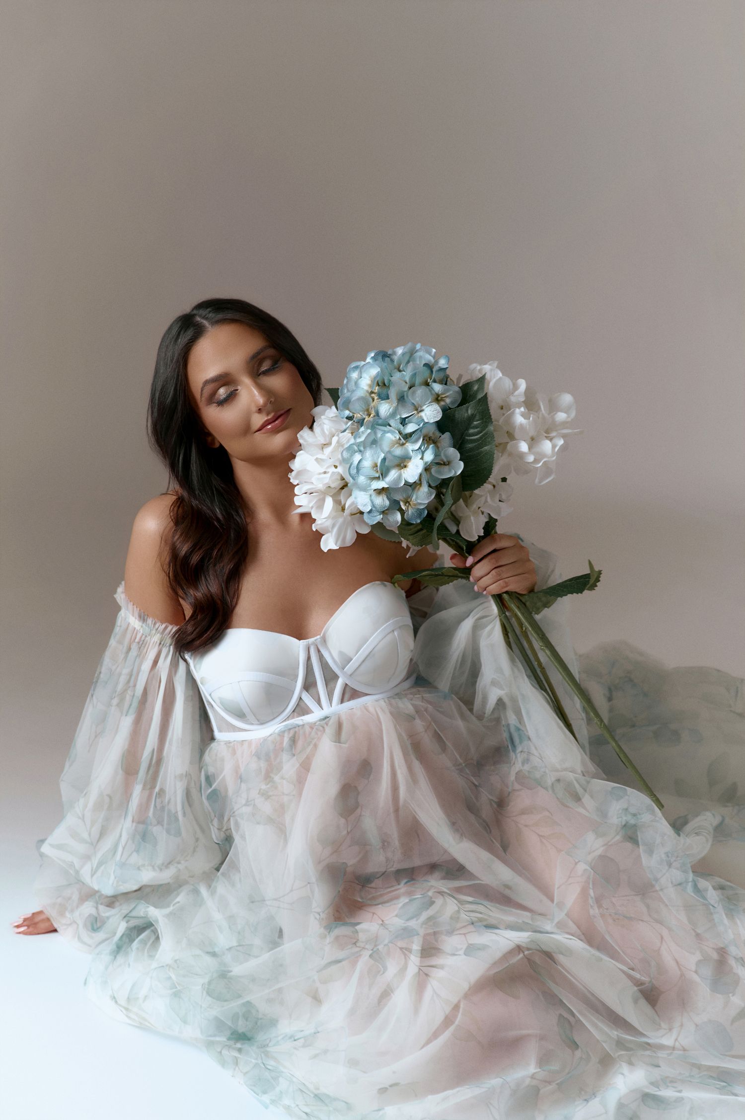 Woman in white corset and sheer floral gown sitting gracefully, holding blue and white hydrangea bouquet.
