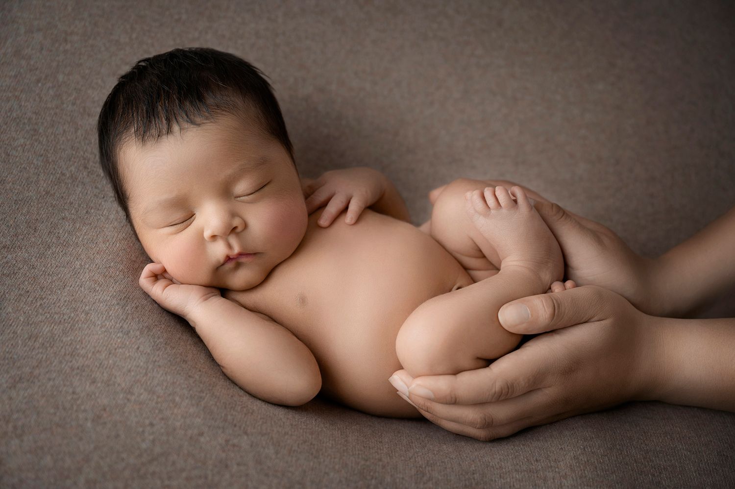 Peaceful newborn baby sleeping on a gray blanket while being gently held by parent hands.
