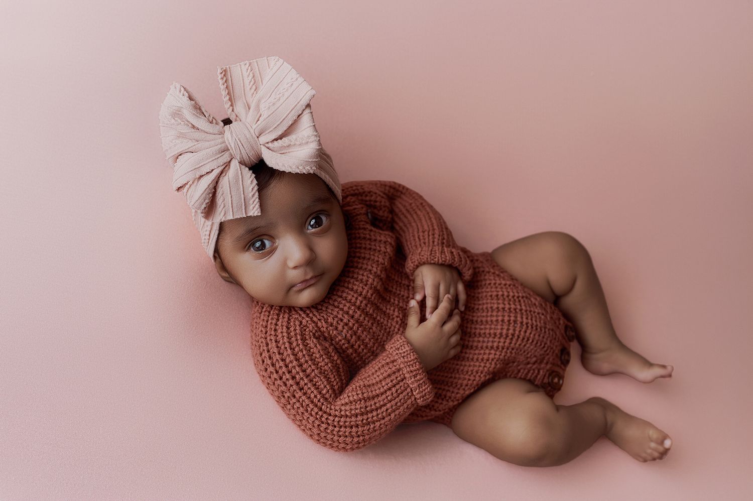 Adorable baby girl in pink knit romper and bow headband poses on pink background.