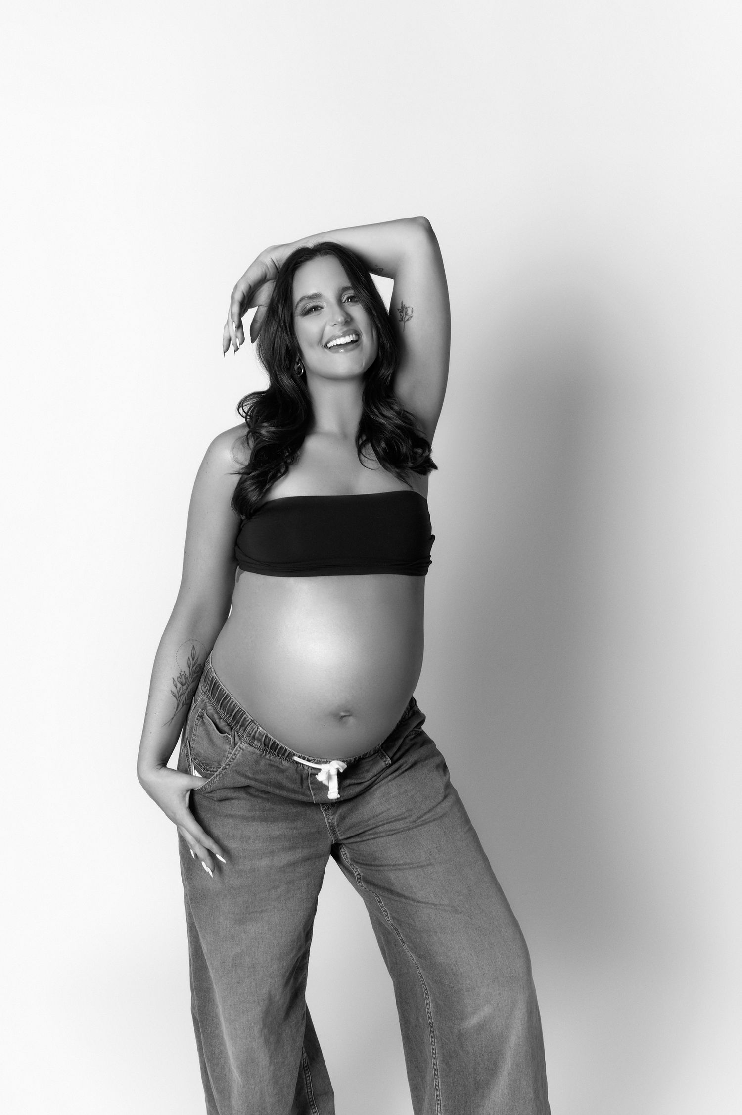Smiling pregnant woman in black tube top and jeans posing with arm raised, black and white maternity photo.