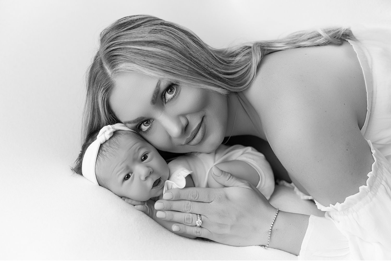 Black and white photo of a smiling mother lying down cheek-to-cheek with her newborn baby girl wearing a white headband.