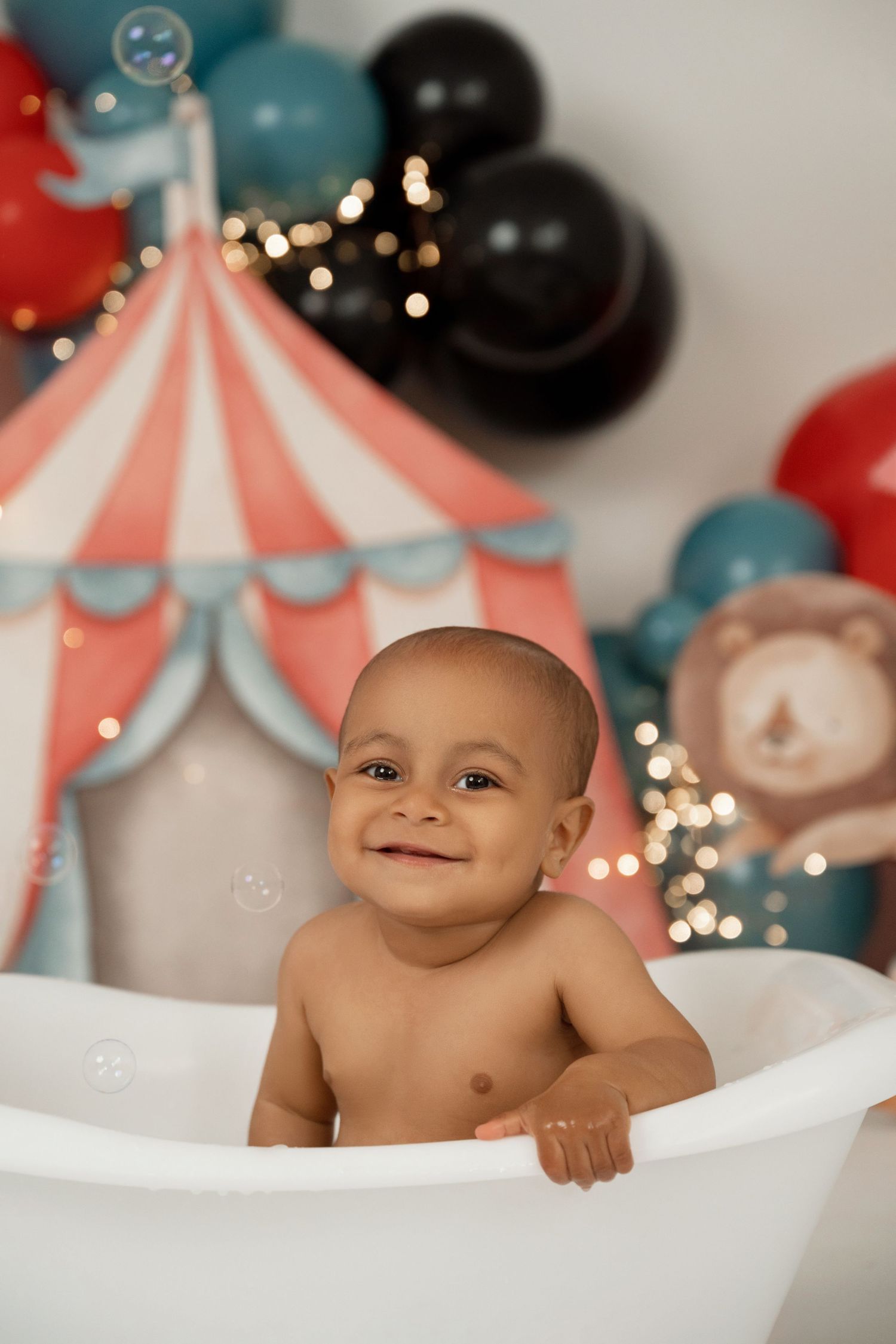 Smiling baby in a white bathtub with a circus-themed backdrop featuring balloons and a colorful tent.