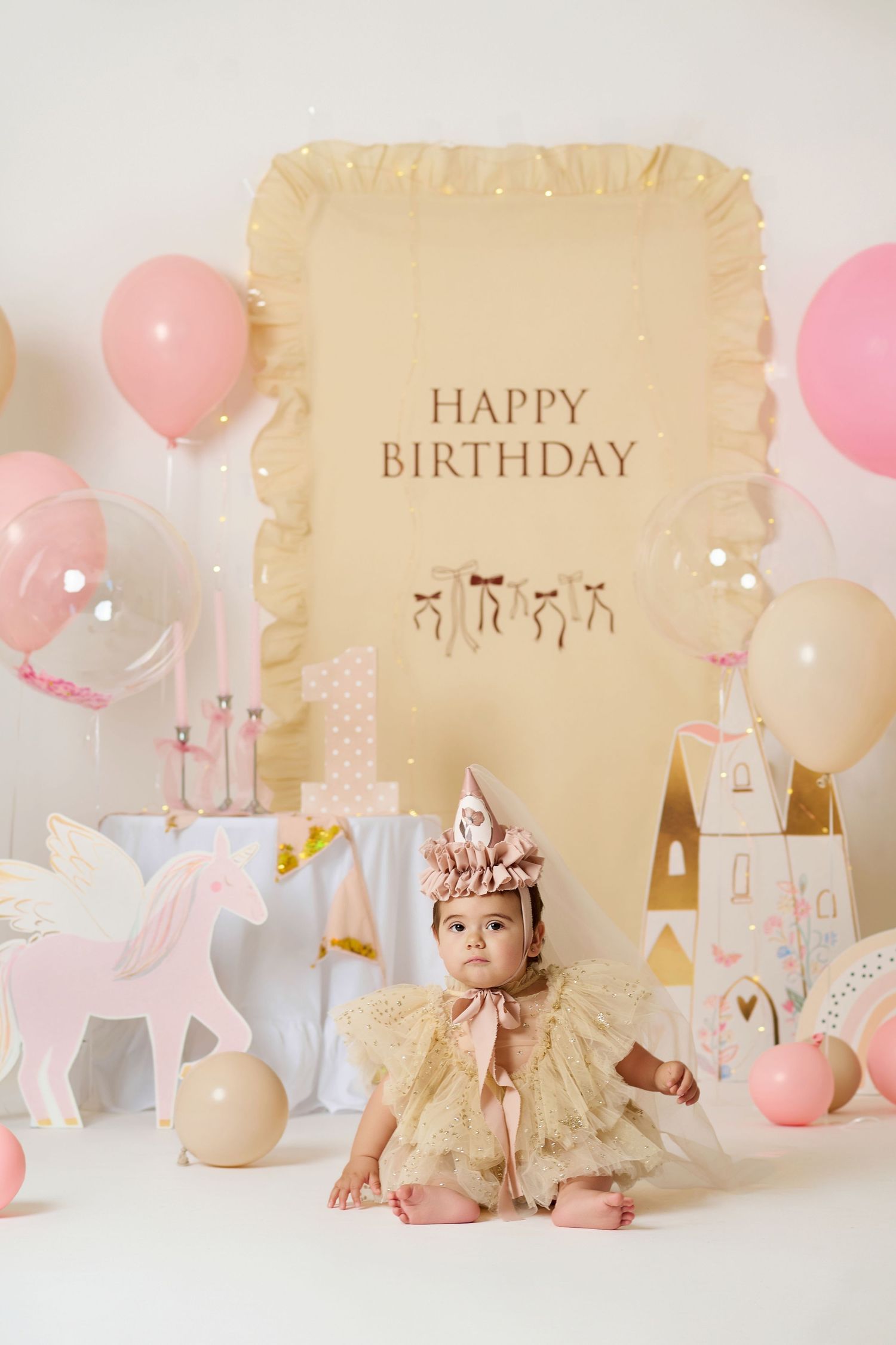 Baby girl in gold dress and crown sits before a Happy Birthday backdrop with pink balloons and unicorn decorations.