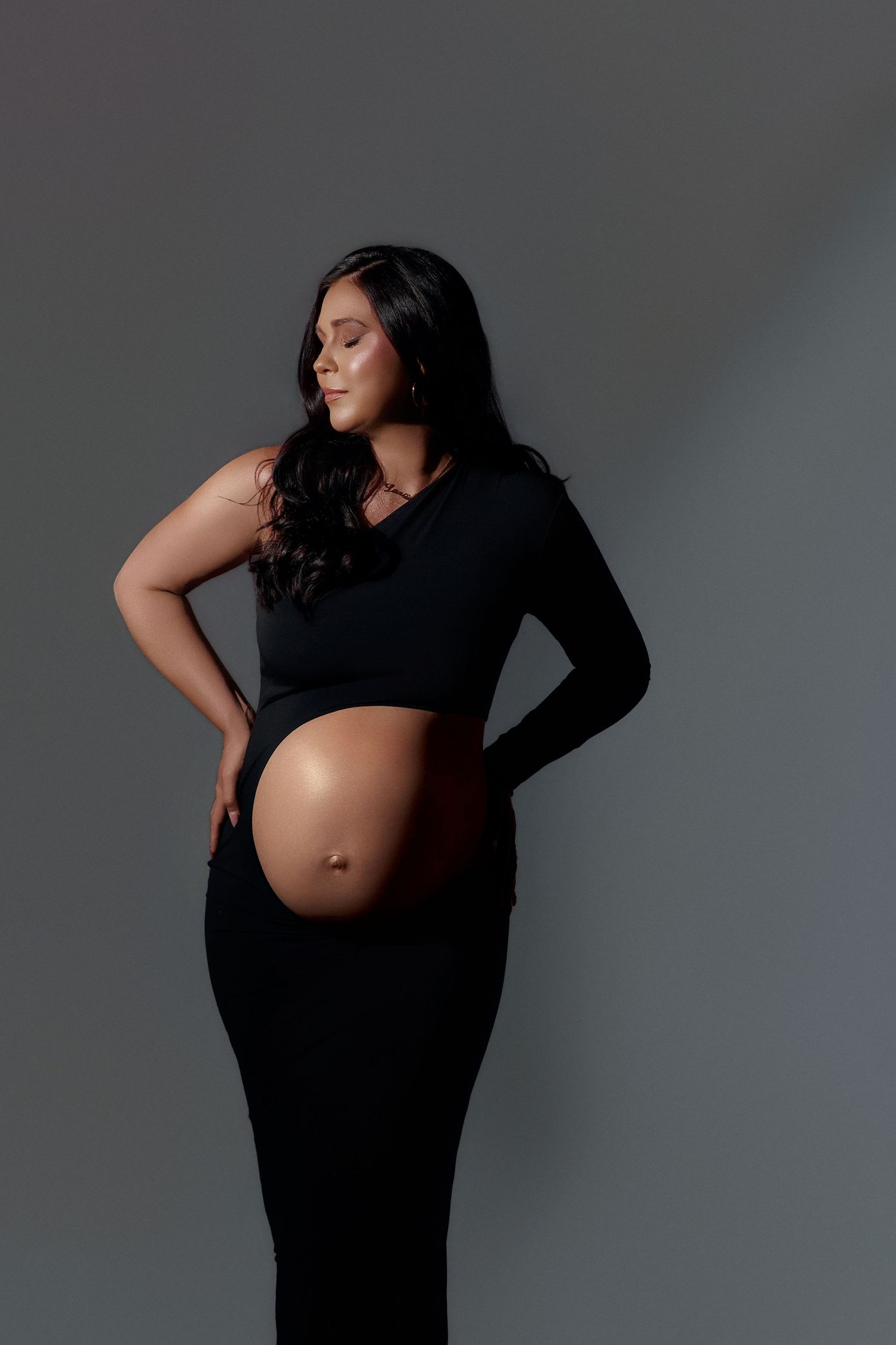 Pregnant woman in black one-shoulder crop top and skirt poses against gray backdrop, cradling baby bump.