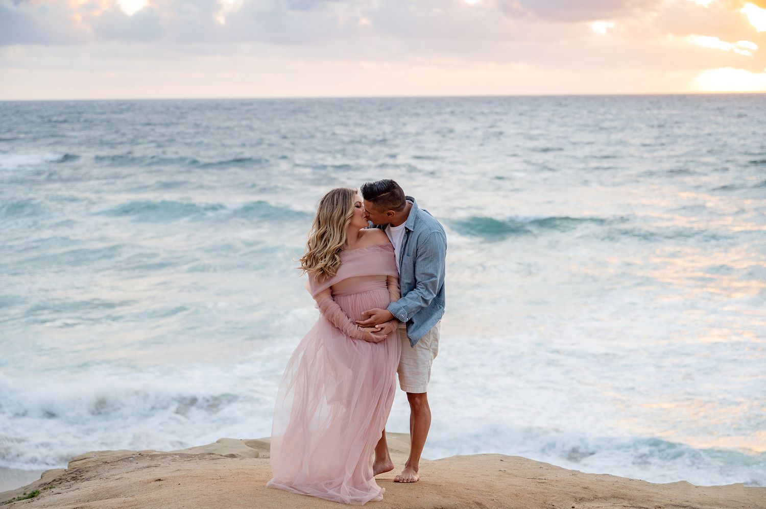 Couple sharing a kiss on rocky beach at sunset, woman in pink maternity gown.