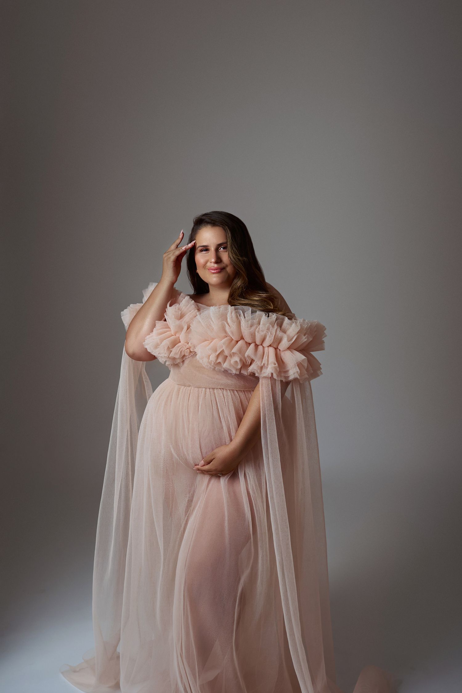 An elegant pregnant woman in a flowing pink ruffled tulle gown poses gracefully in a soft gray studio setting.