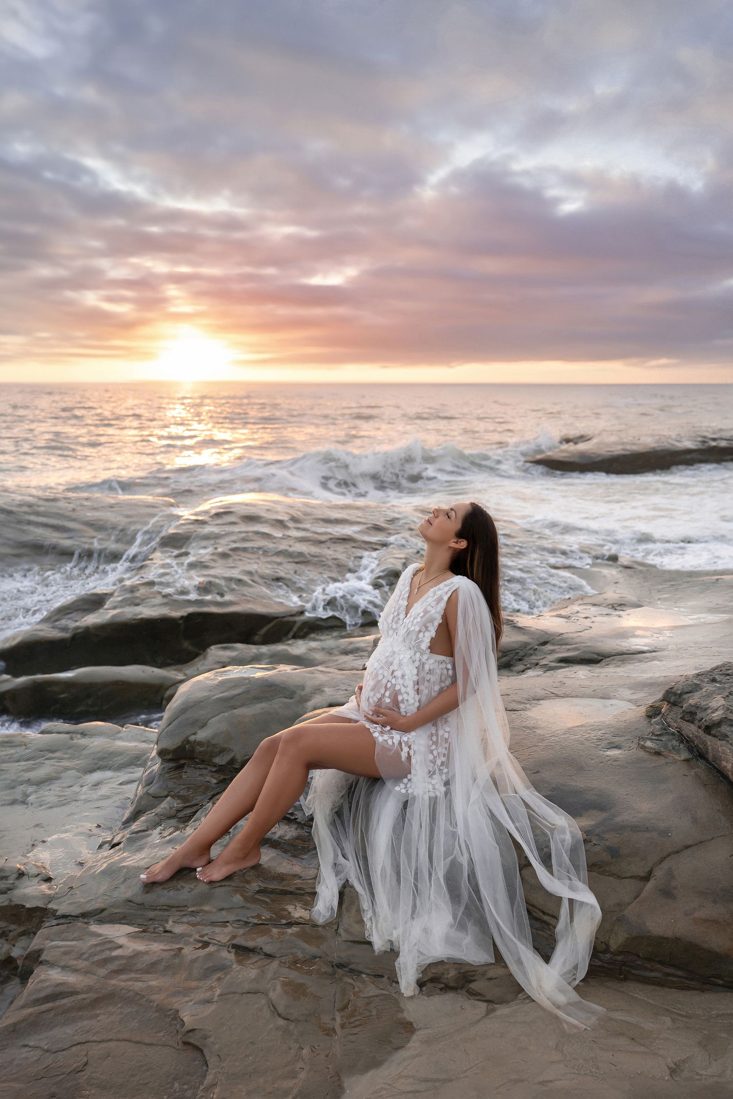 Pregnant woman in white lace and tulle gown sits on coastal rocks gazing upward during dramatic sunset.