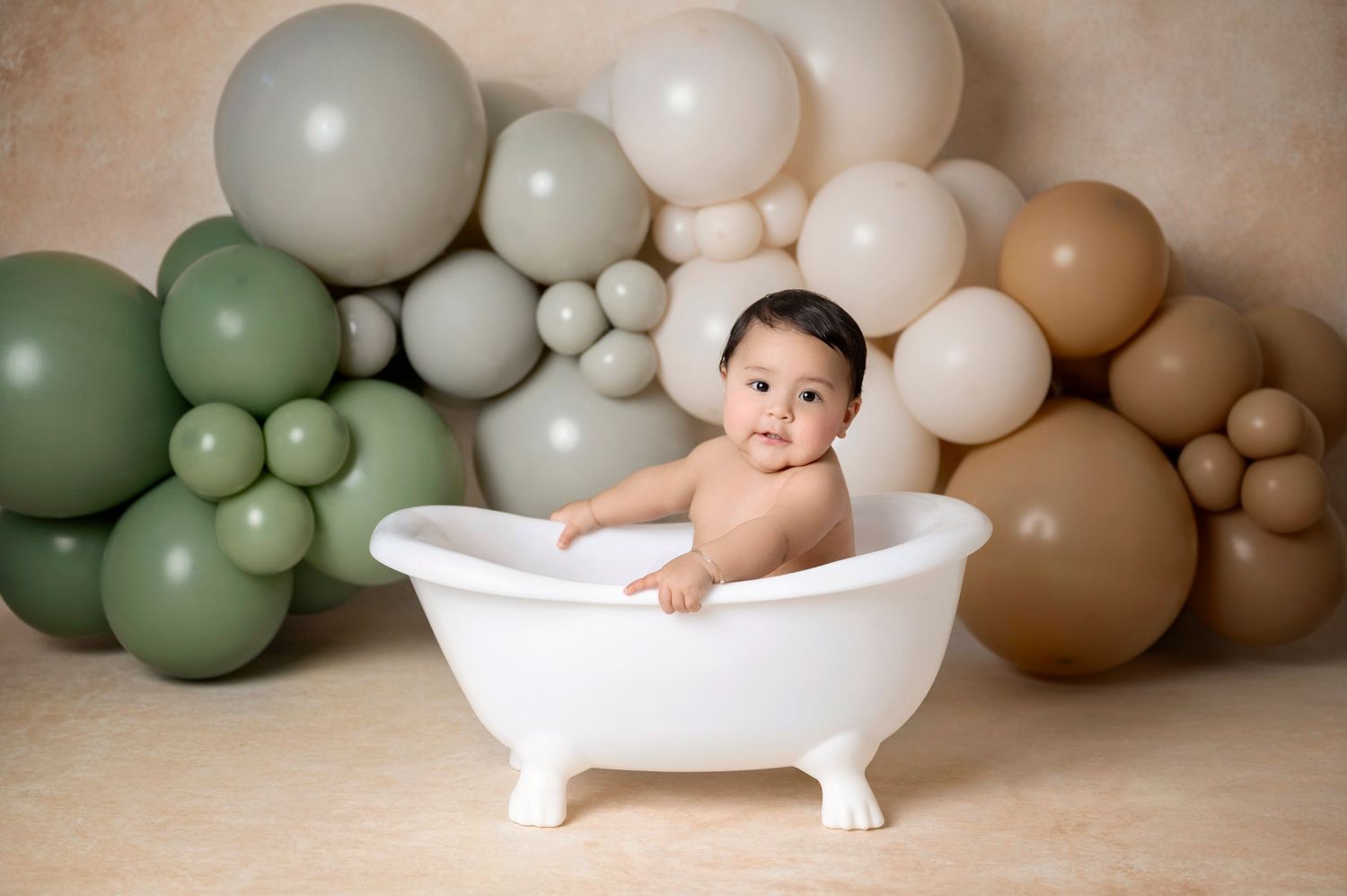 Baby sitting in a white clawfoot tub surrounded by sage green, beige, and tan balloon garland backdrop.