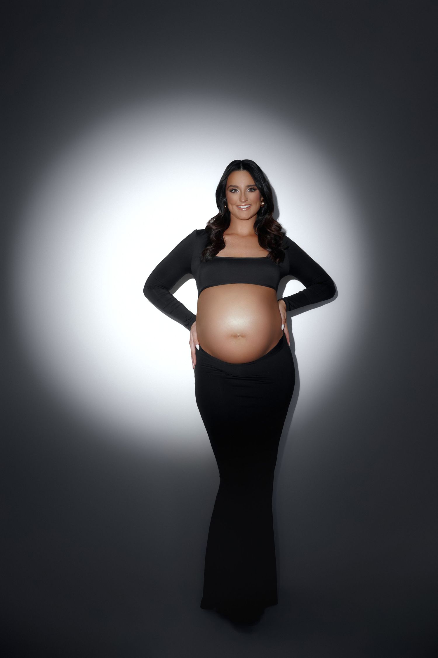 Smiling pregnant woman in black crop top and maxi skirt posing with hands on hips against glowing backdrop.