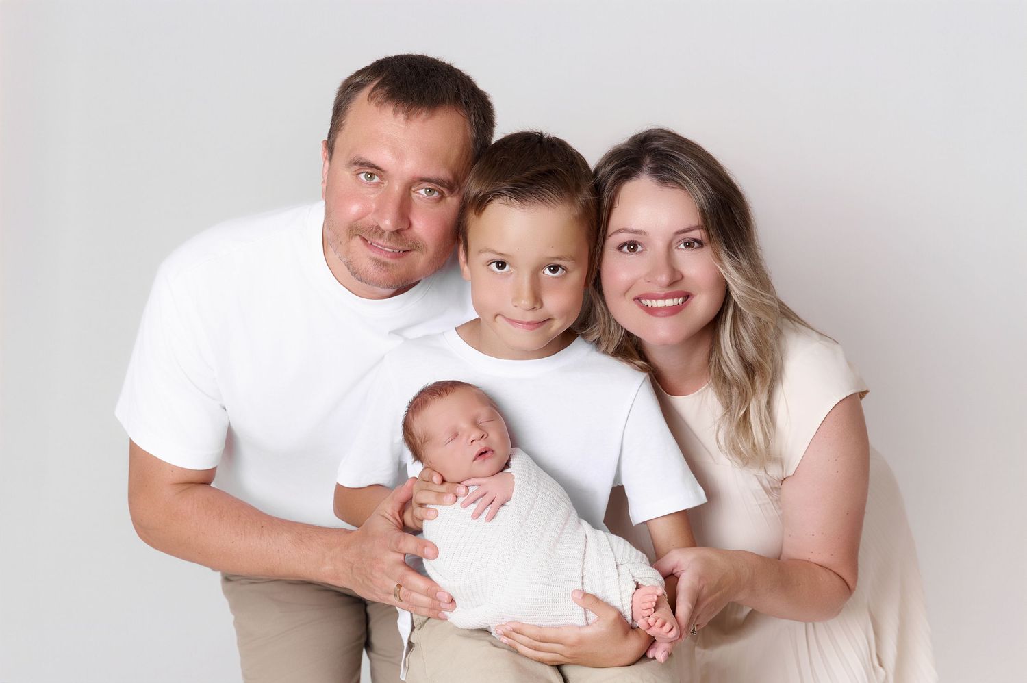 Happy family of four posing with newborn baby, parents and young boy in white shirts on light background.