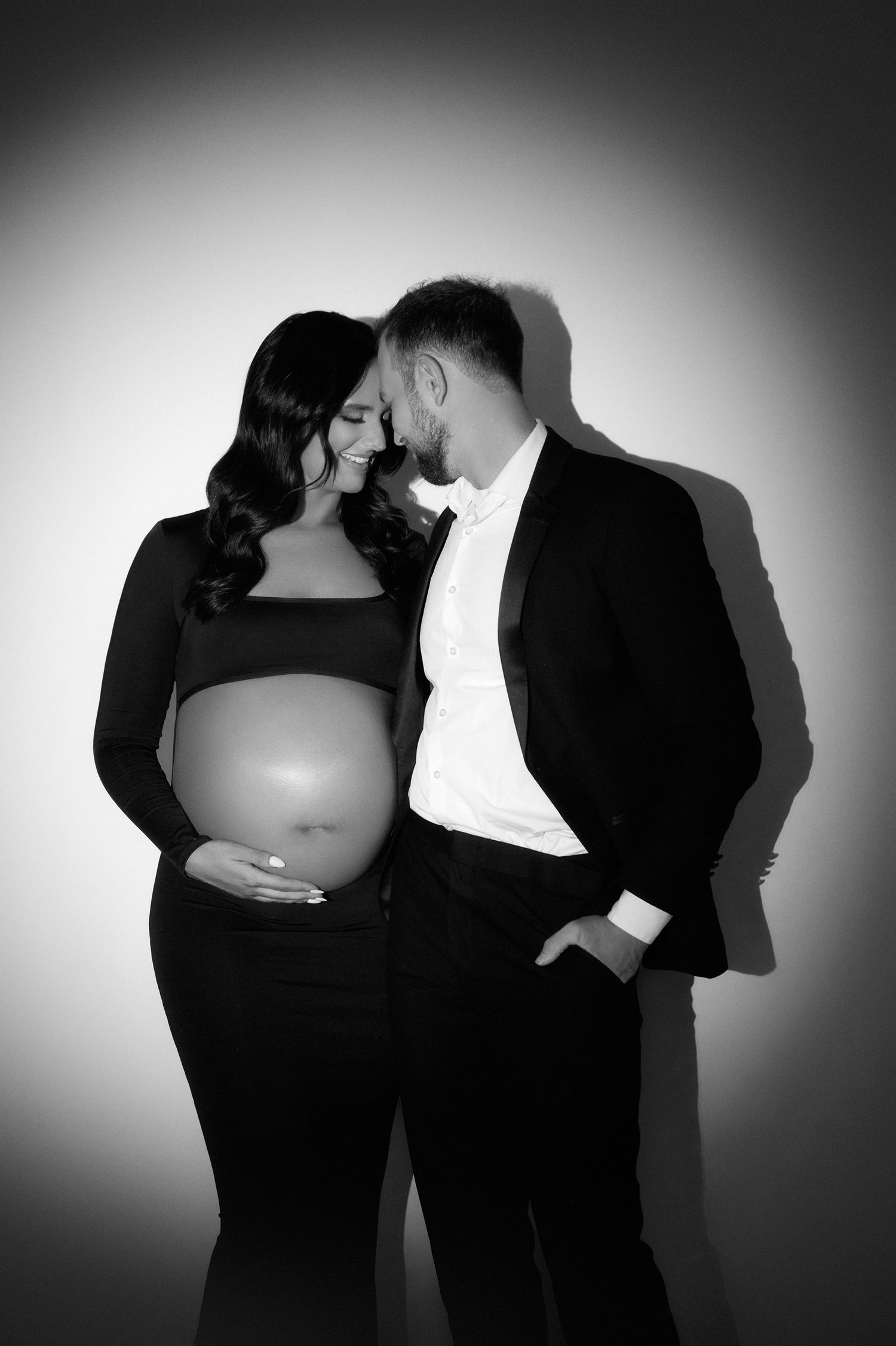 Black and white maternity photo of a couple in formal wear, woman cradling baby bump, man leaning in closely.