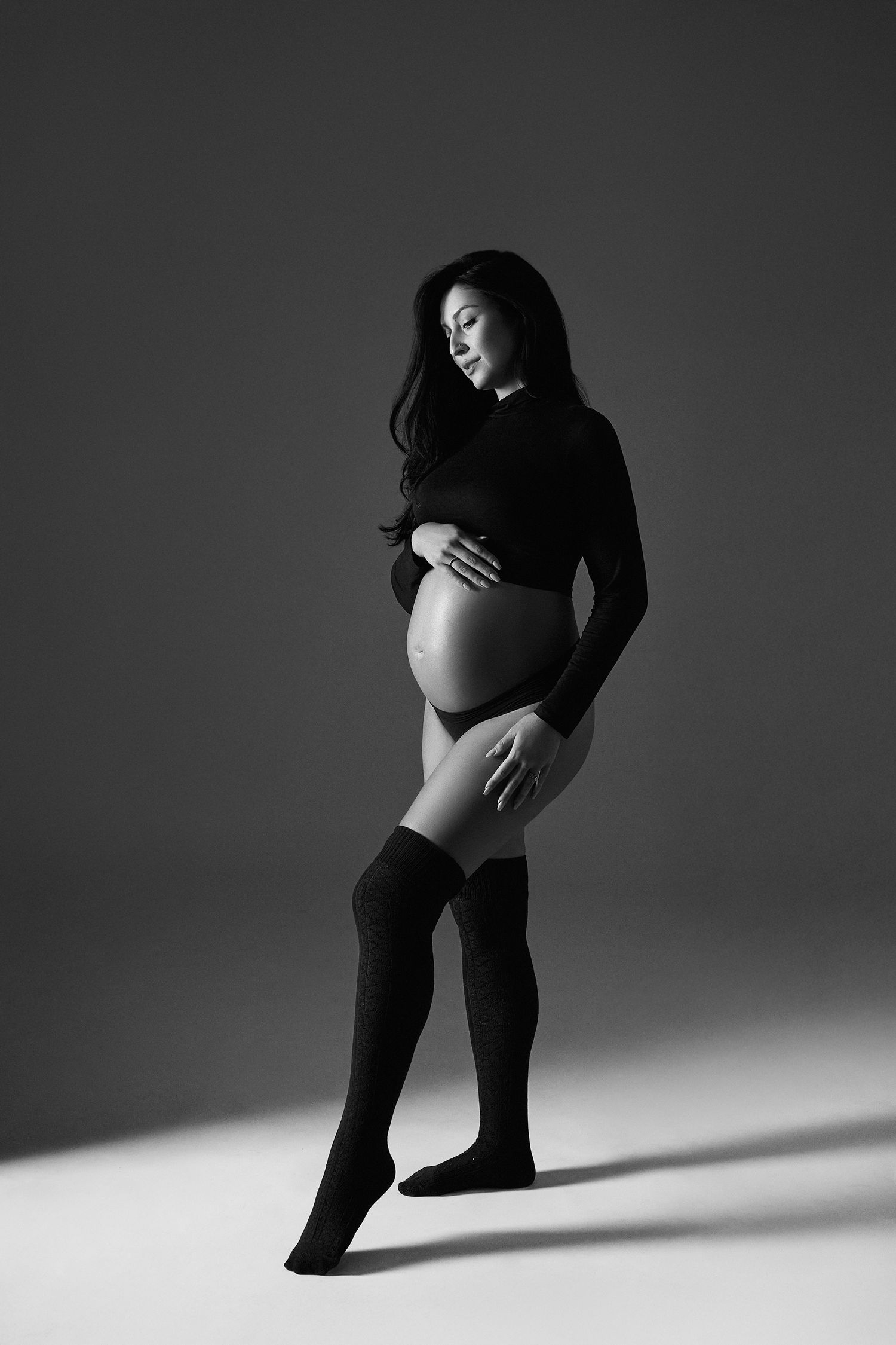 Black and white photo of pregnant woman in black turtleneck and knee-high socks cradling her baby bump.