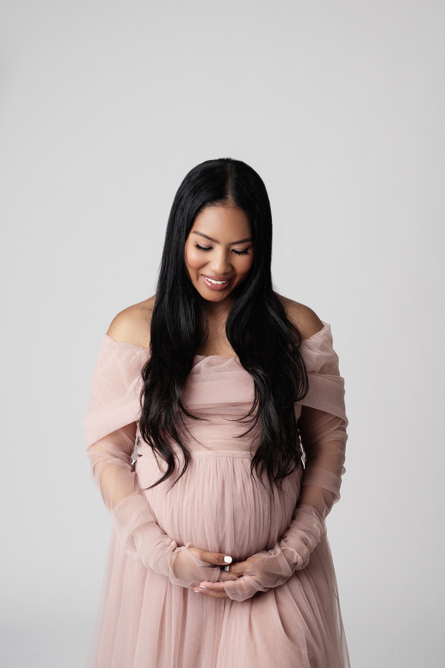 Smiling pregnant woman in blush pink off-shoulder tulle gown holds baby bump in white studio.