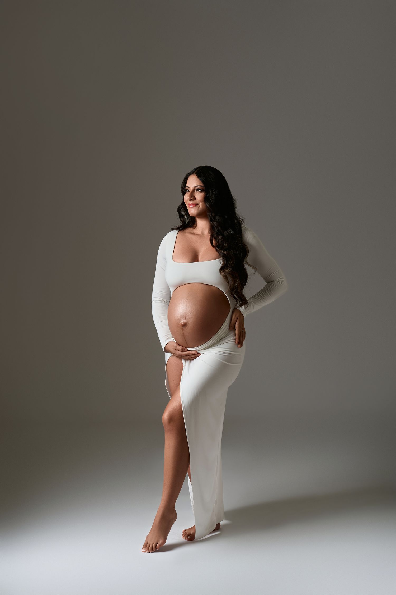 Pregnant woman in elegant white two-piece maternity gown with side slit, holding baby bump in a studio portrait.