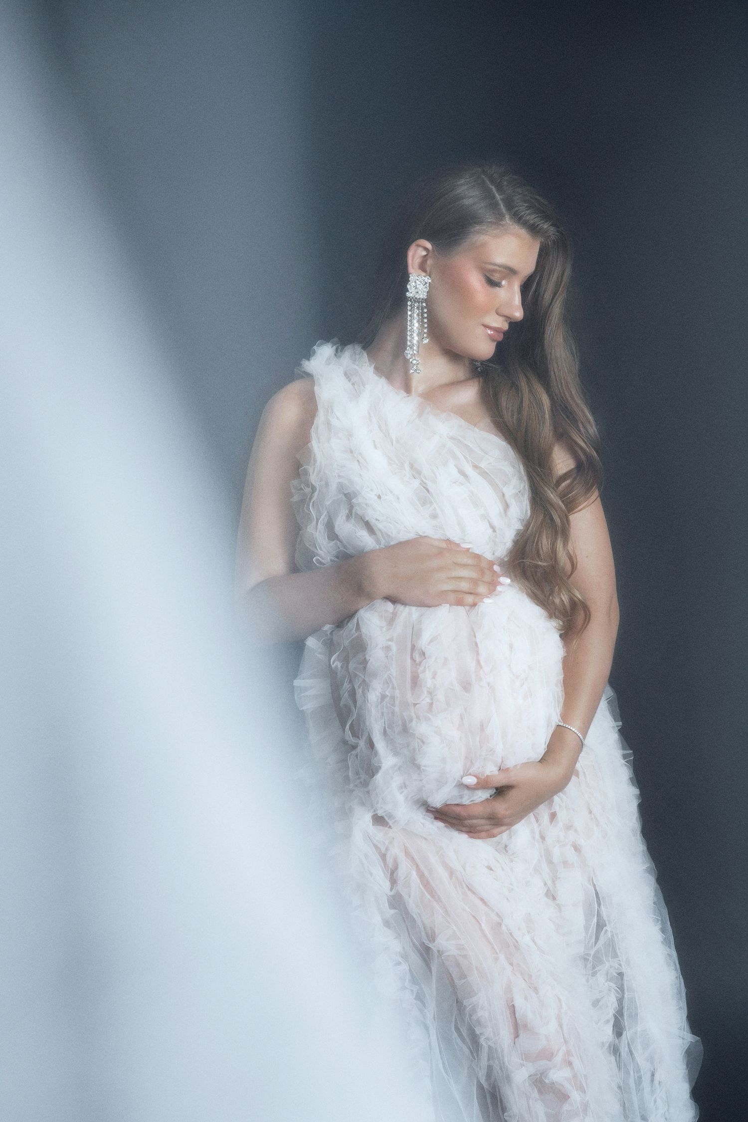 Elegant pregnant woman in white feathered dress cradling baby bump, long wavy hair, chandelier earrings, studio portrait.
