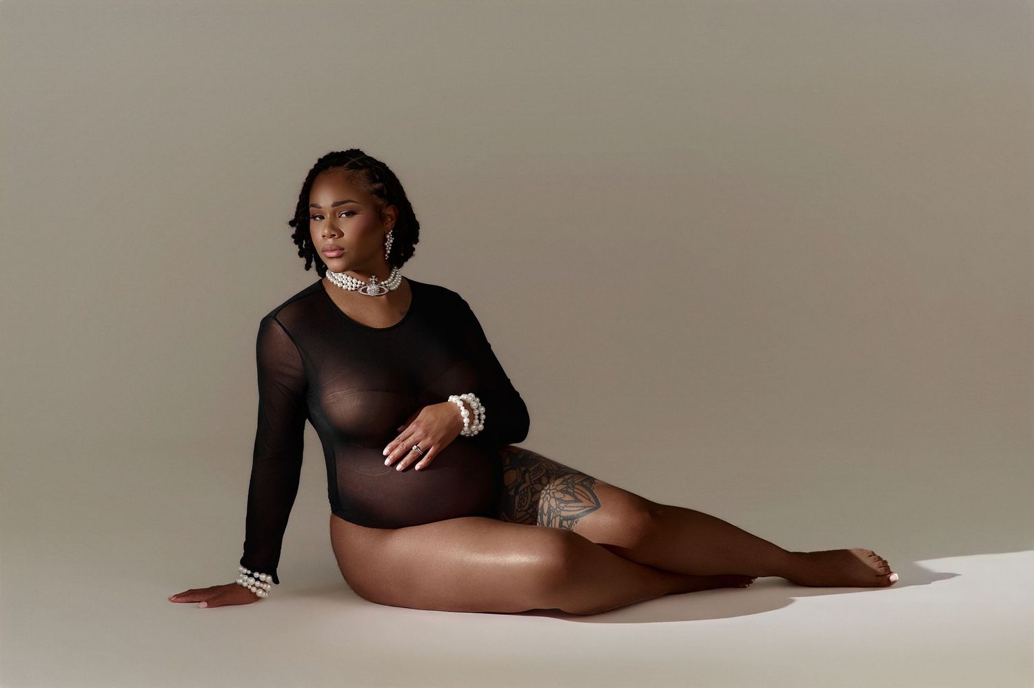 Woman in black sheer bodysuit posing gracefully on floor against neutral background, wearing pearl jewelry.