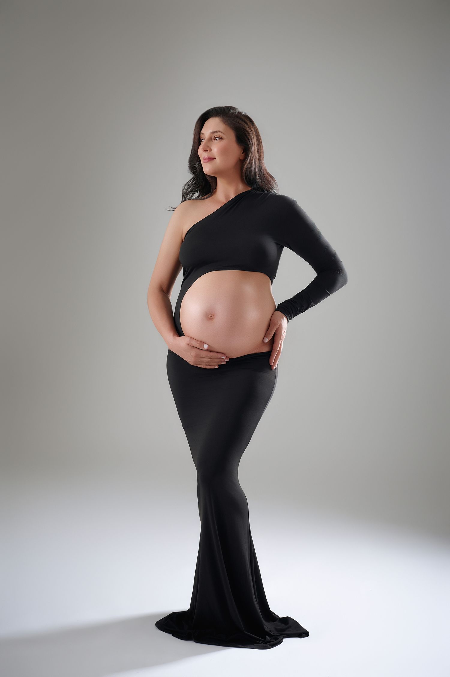 Pregnant woman in black one-shoulder crop top and maxi skirt cradling her baby bump in San Diego studio maternity photo.