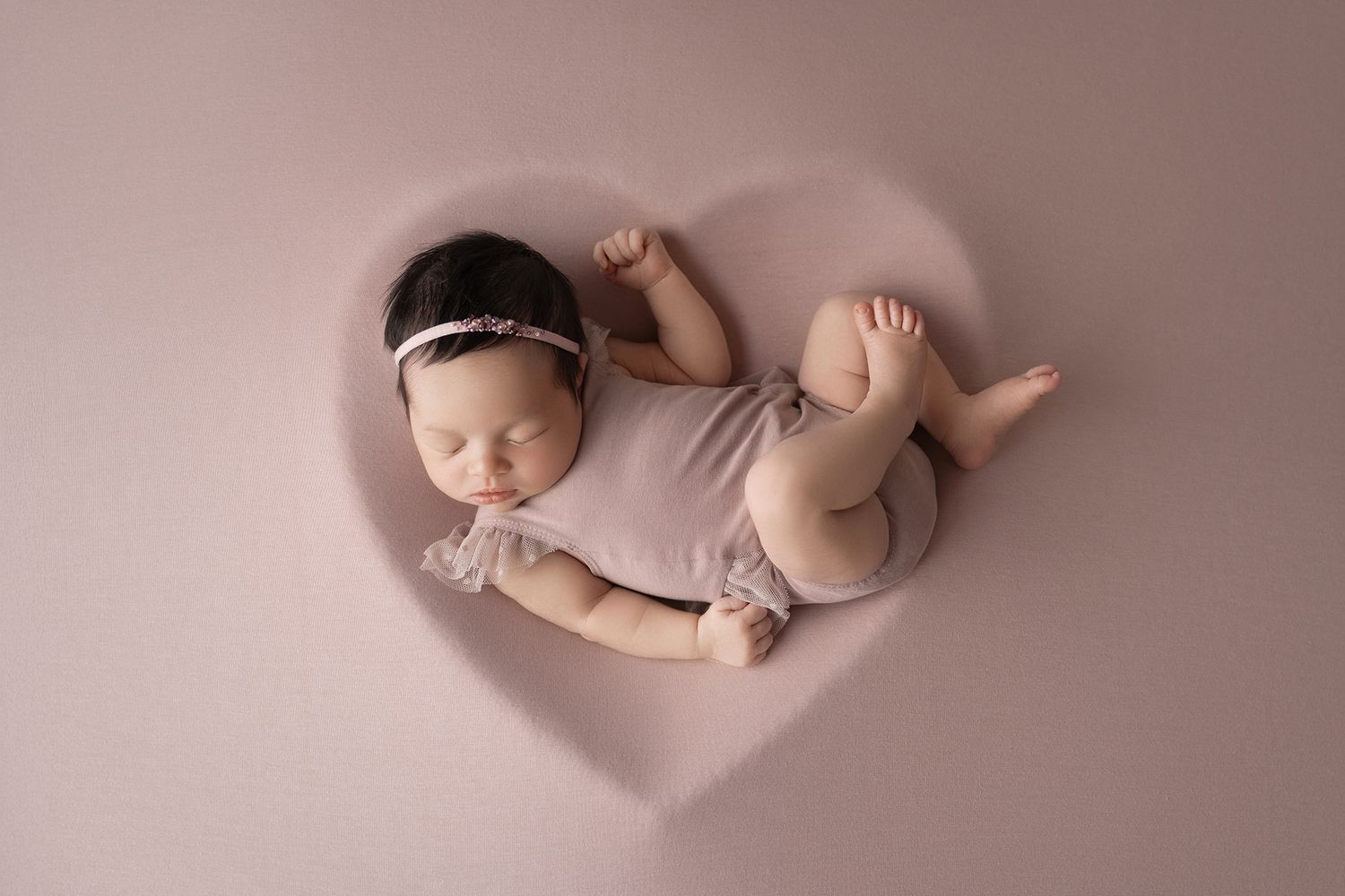 Newborn baby girl sleeping peacefully in a heart-shaped prop, wearing a mauve romper and floral headband.