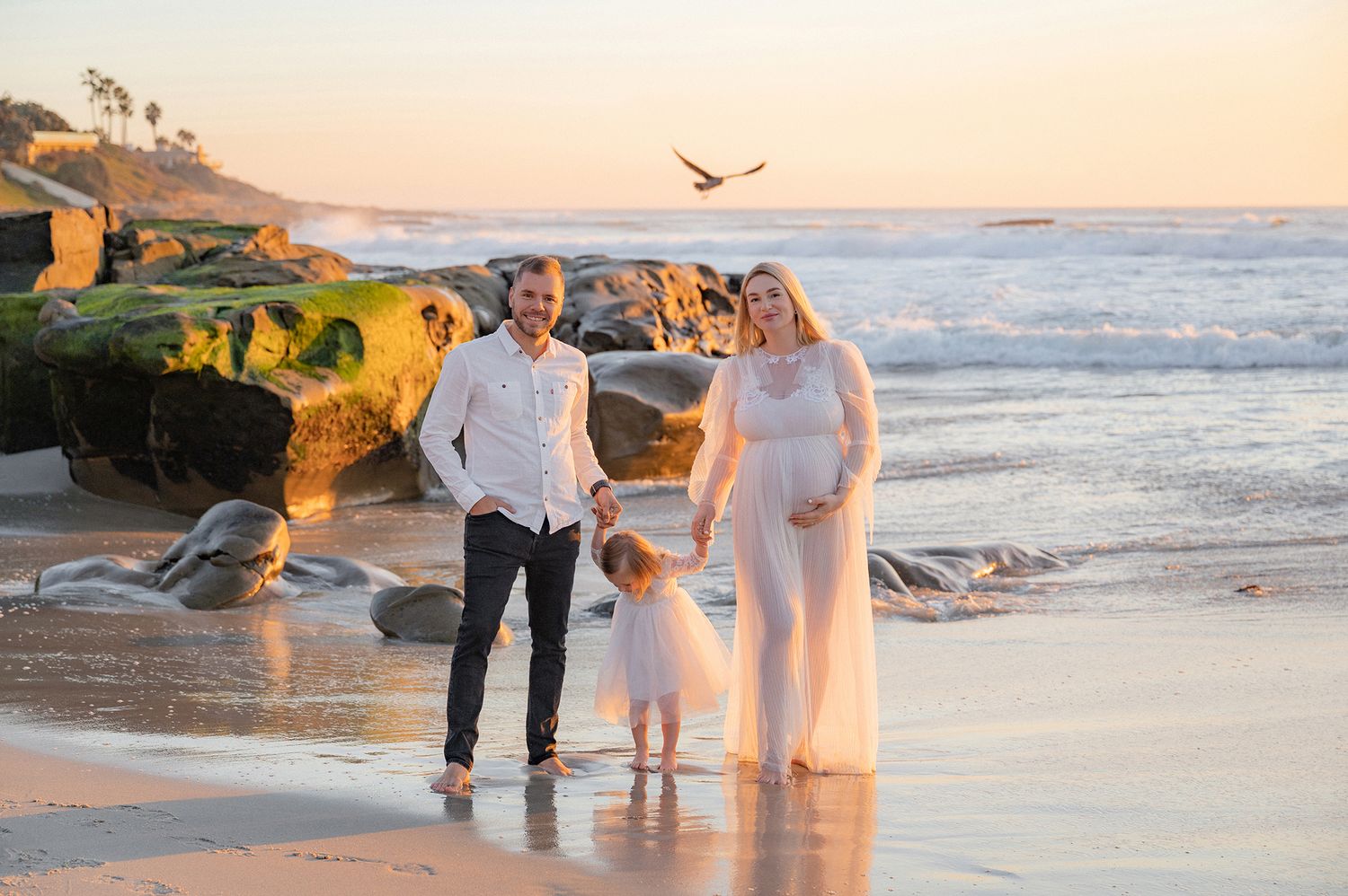 Family maternity photo on sandy beach at sunset with parents and young daughter holding hands.
