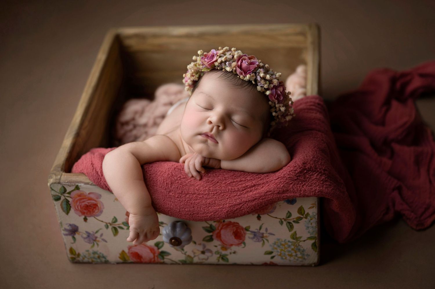 Sleeping newborn baby girl wearing a floral crown nestled in a wooden box with pink wraps for a studio photo session.