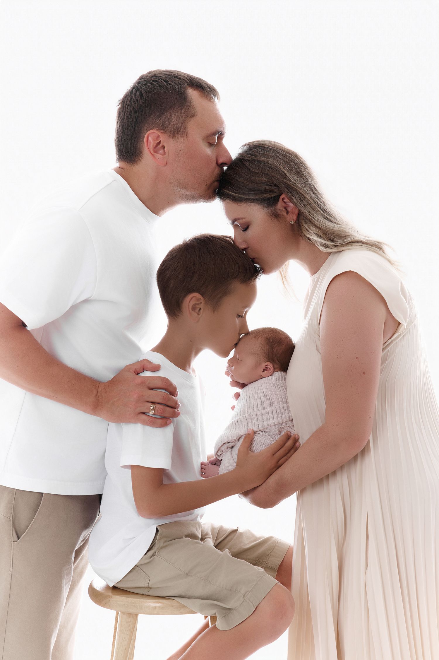 Family of four in white outfits sharing tender kisses with newborn baby in a bright studio portrait.