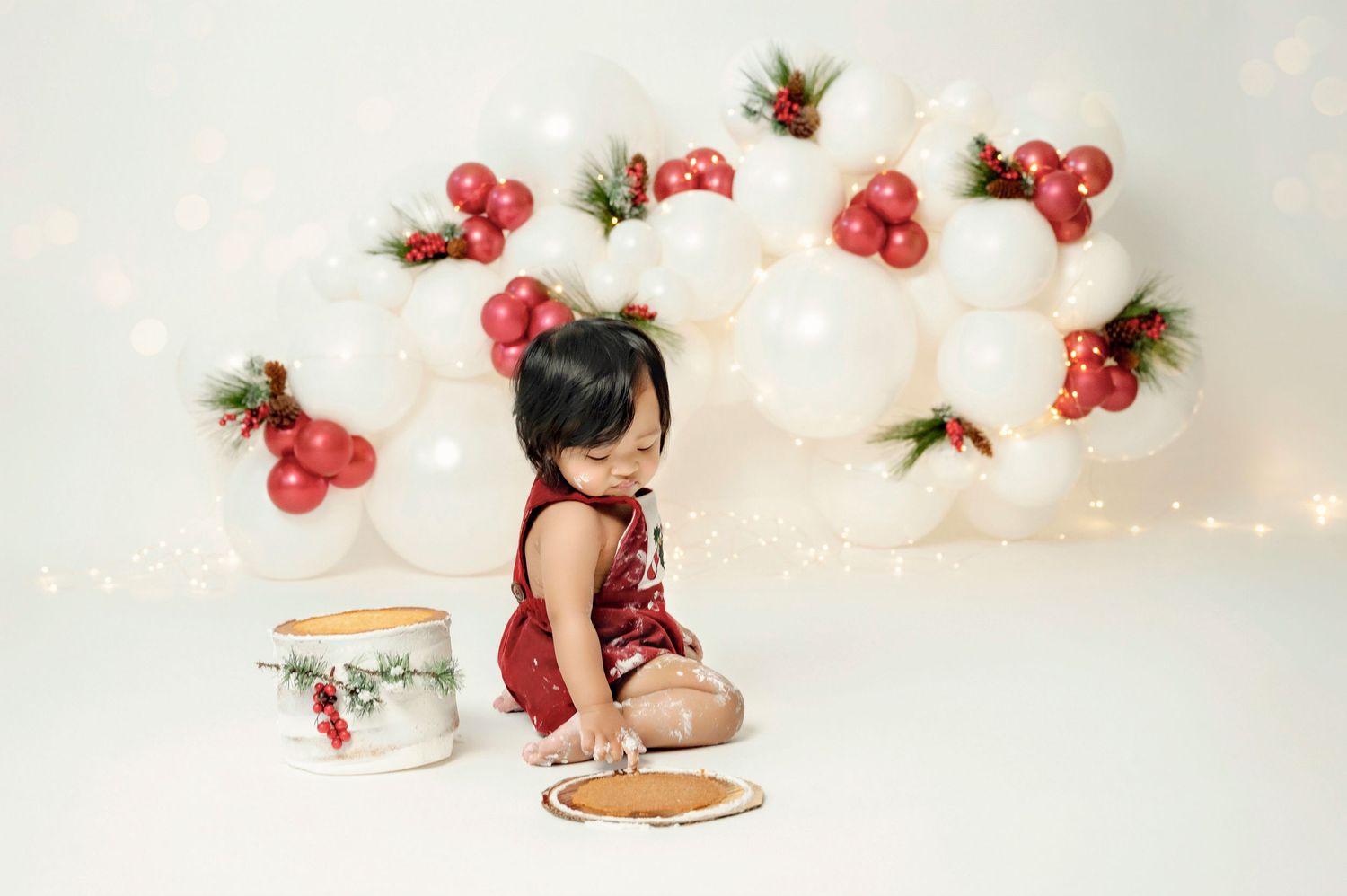 Baby girl in red romper smashes cake at Christmas-themed photo session with balloon backdrop.