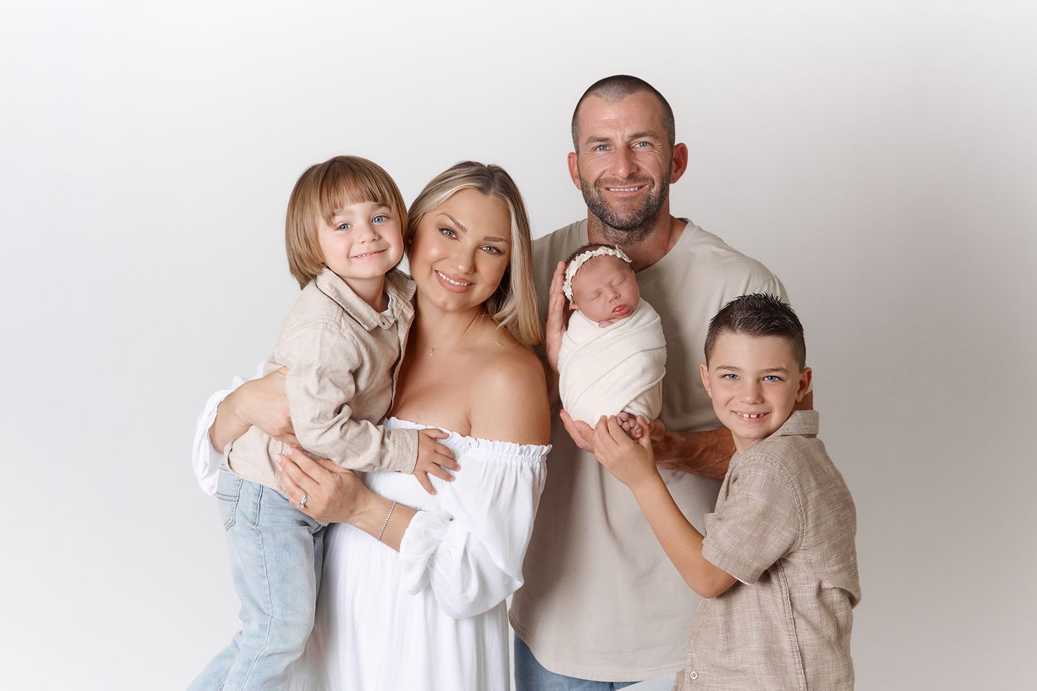 Family of five posing for newborn portrait, parents with two boys and swaddled baby, white studio background.