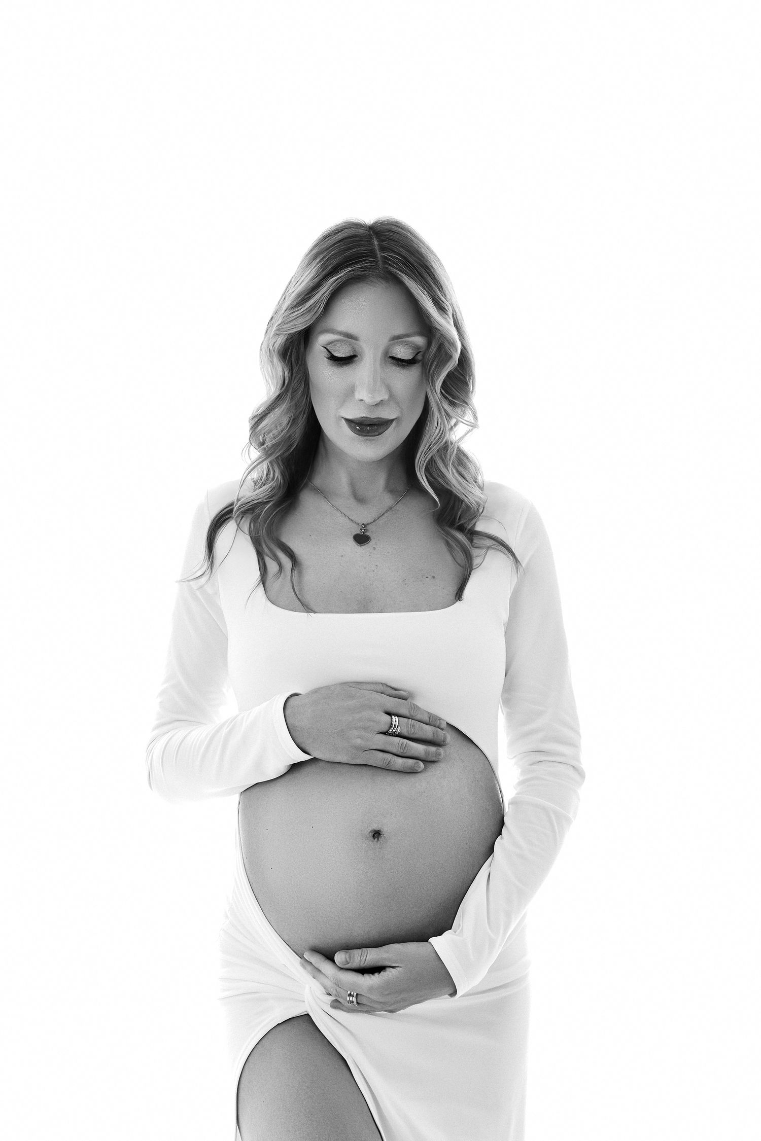 Black and white portrait of a pregnant woman in a white dress cradling her baby bump, looking down tenderly.