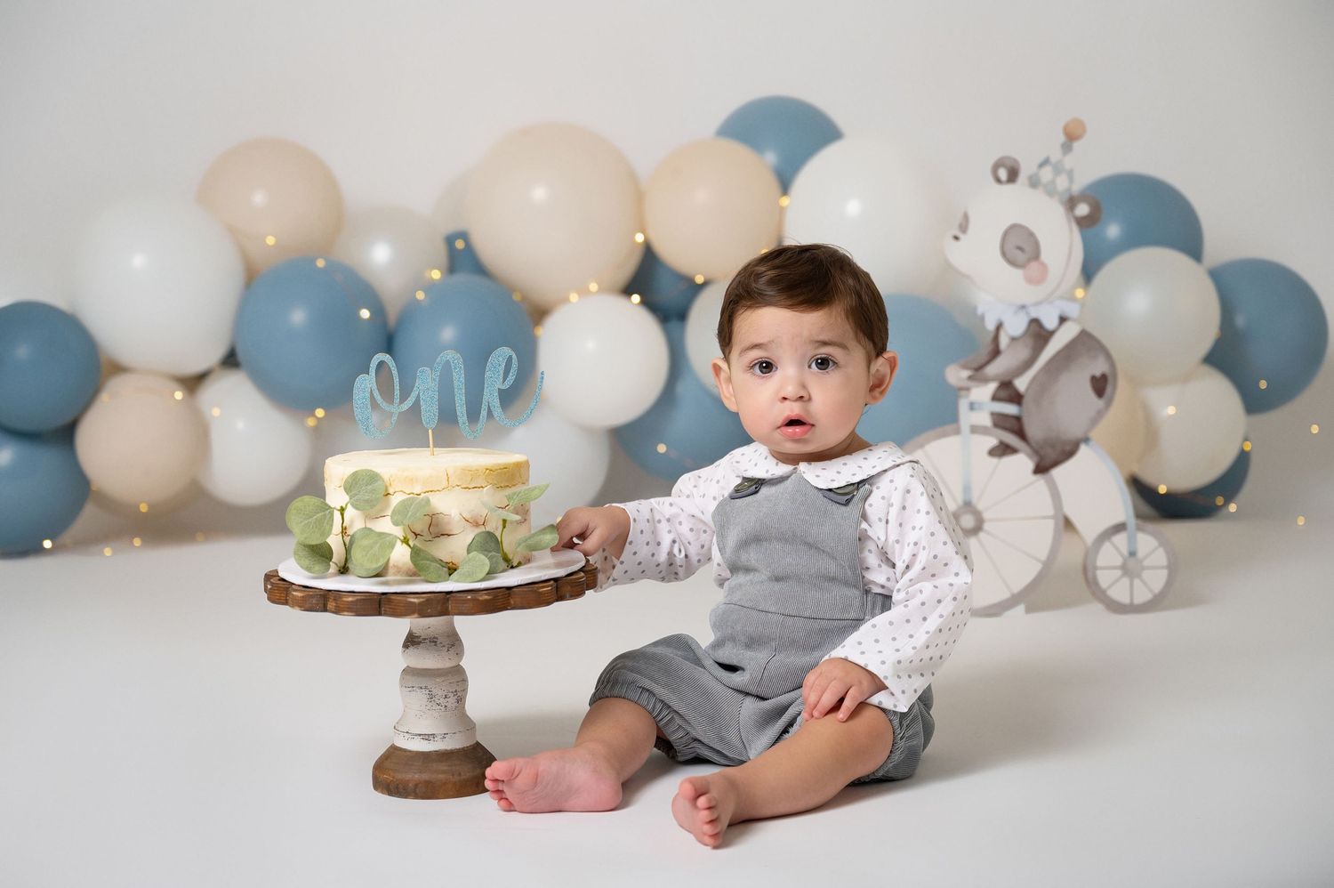 Baby boy in gray vest sits beside a cake smash cake with balloons during a first birthday photo session.
