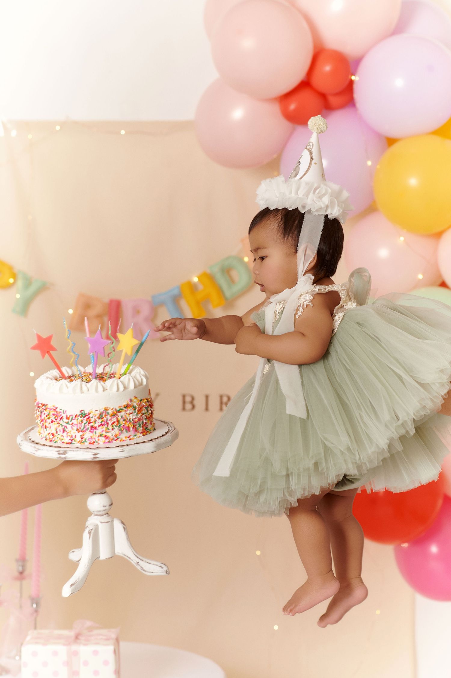 Baby girl in green tutu dress reaching for birthday cake with colorful candles, balloons and party decorations.