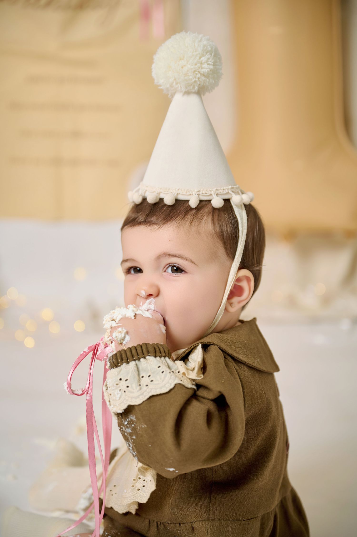 A baby in a white party hat and olive dress eats cake during a first birthday cake smash photo session.