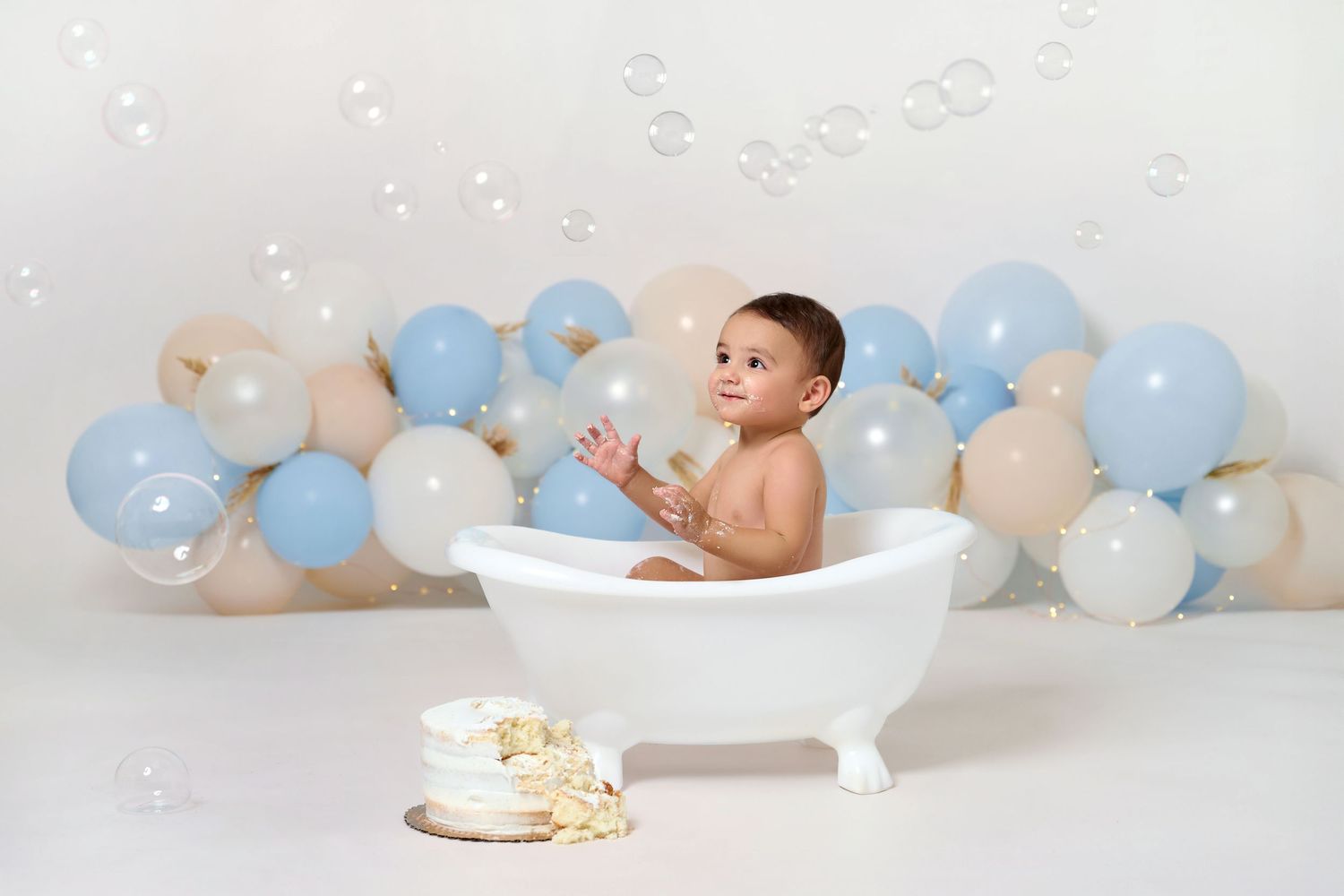 Baby in white bathtub with blue and beige balloons, bubbles floating, smash cake nearby, first birthday photo session.
