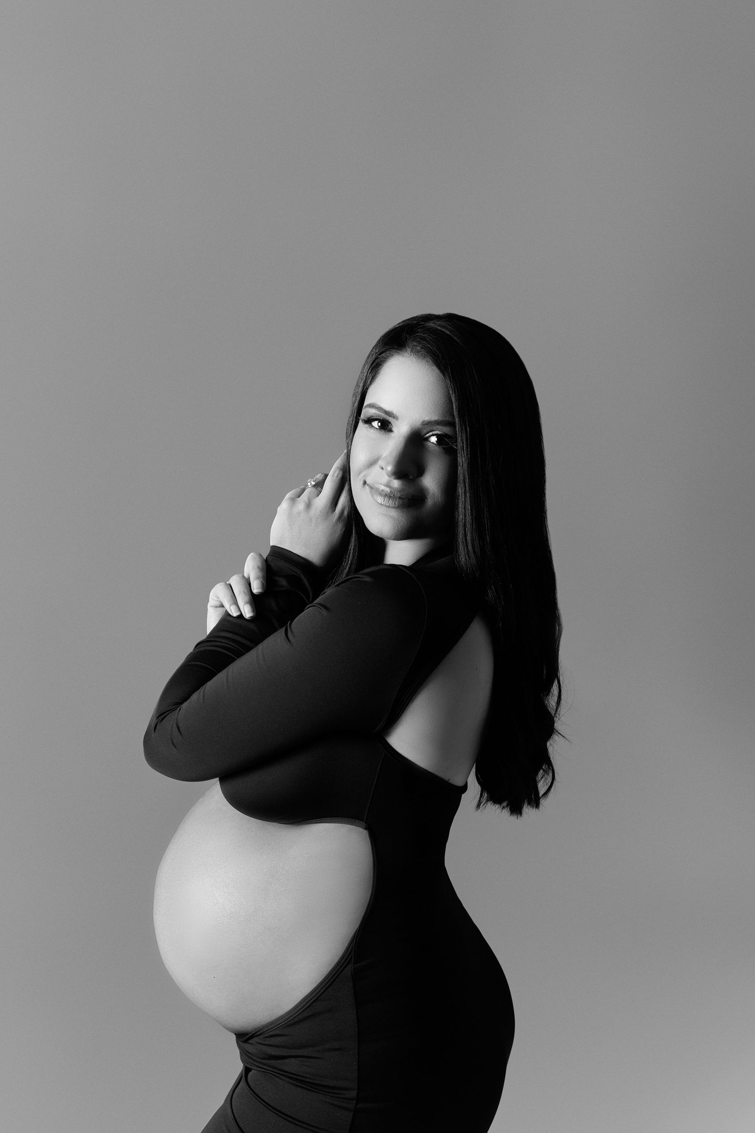 Black and white maternity portrait of pregnant woman in backless dress cradling her baby bump.