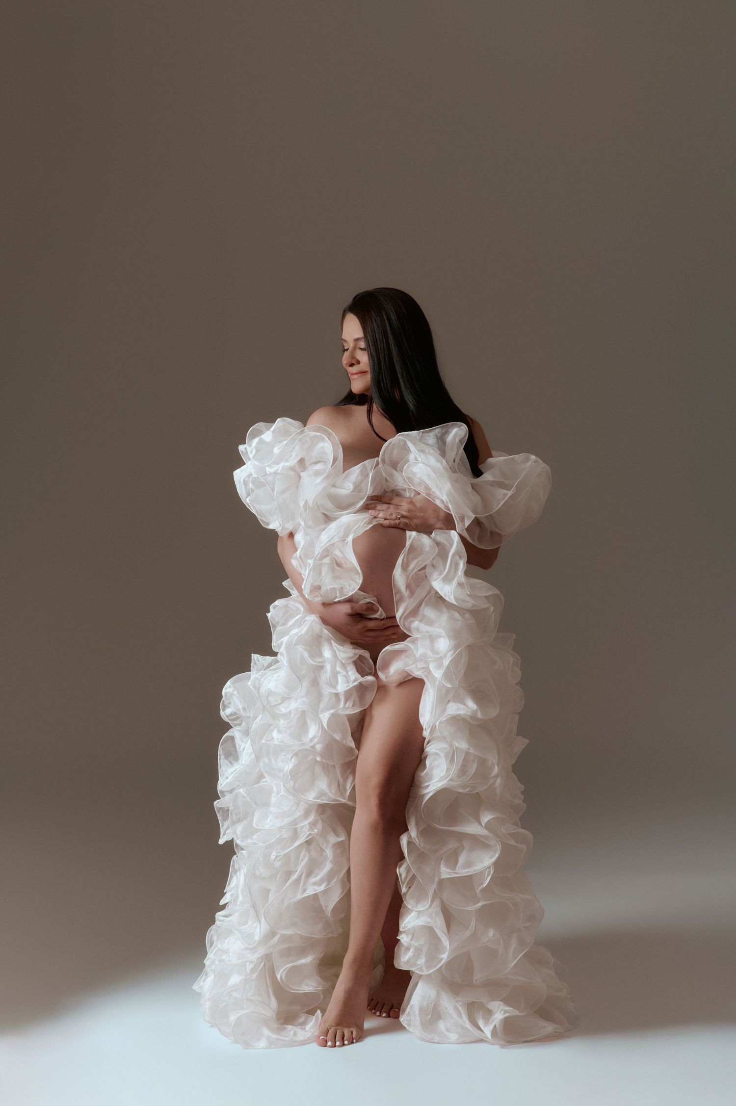 Pregnant woman in a flowing white ruffled maternity gown poses gracefully in a studio setting.