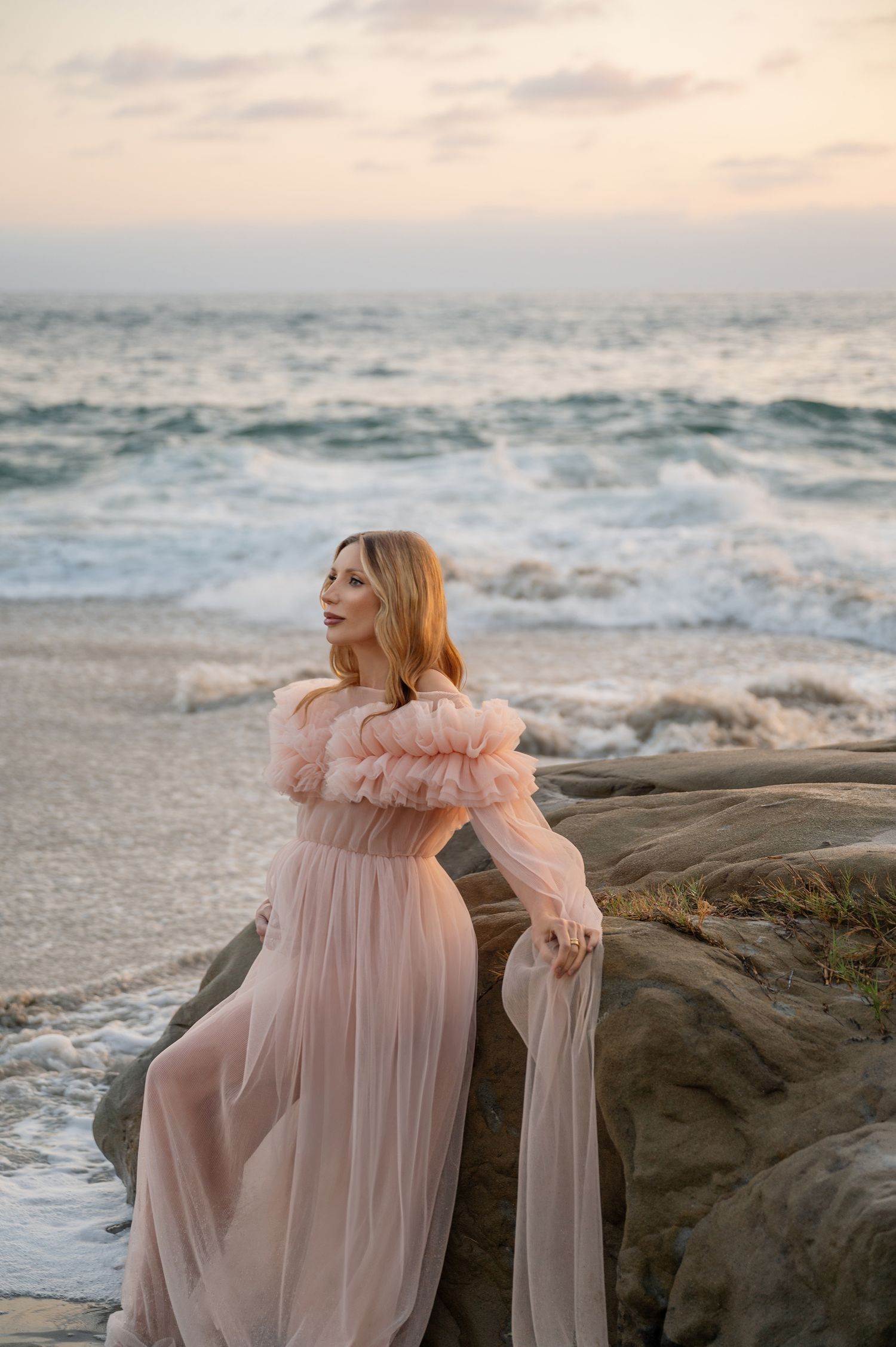 Pregnant woman in pink ruffled off-shoulder gown leaning on coastal rocks at dramatic sunset.