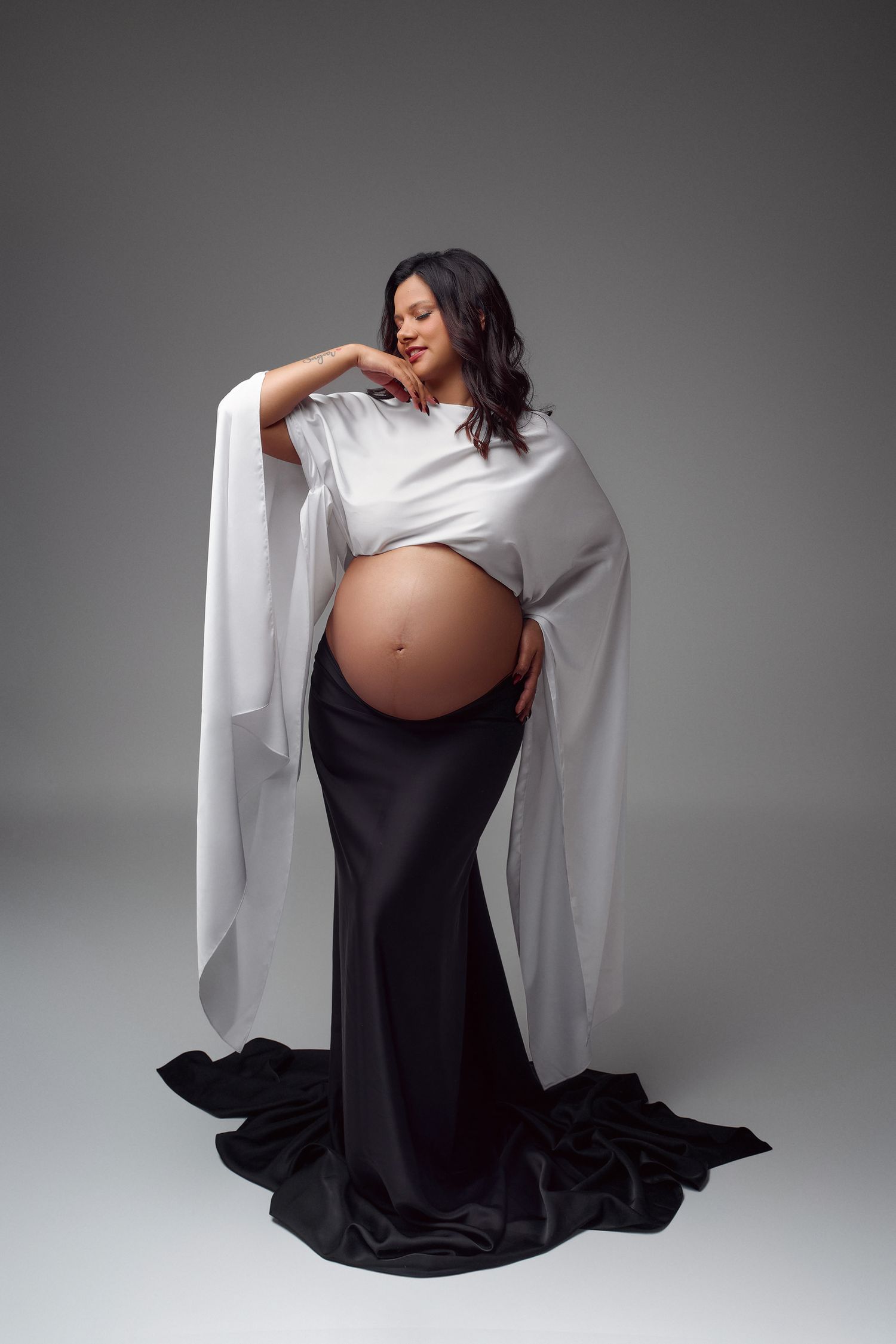 Pregnant woman in white draped top and black satin skirt posing gracefully in studio maternity photo shoot.