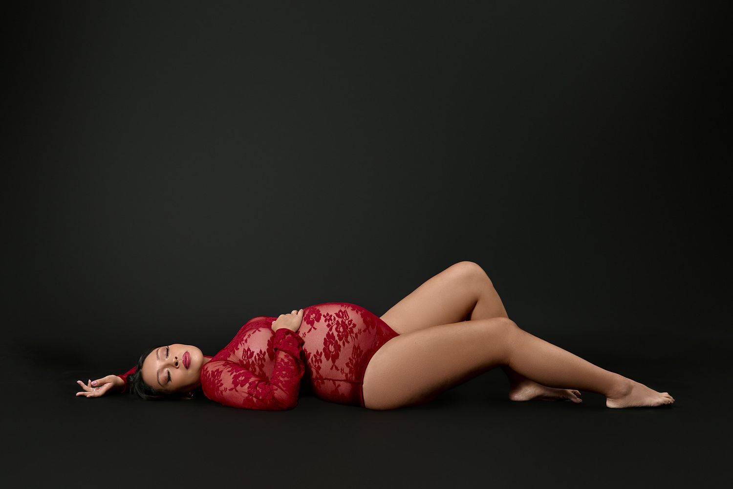 Woman in red lace bodysuit posing gracefully on dark studio floor.