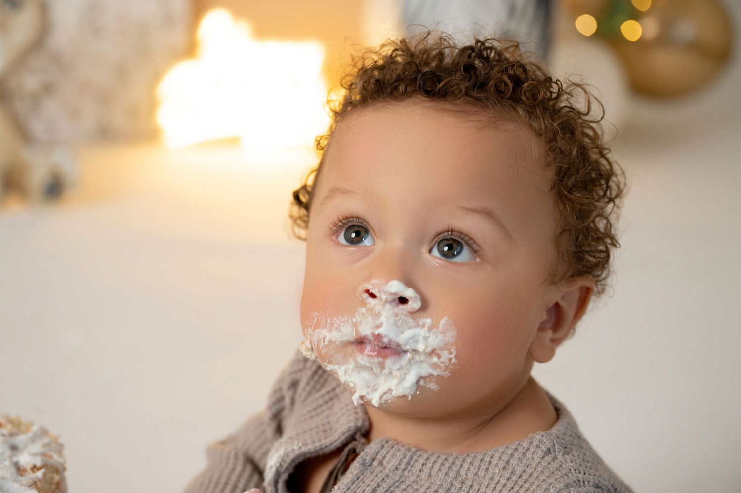 Adorable baby with curly hair and blue eyes enjoying cake smash with white frosting on face.
