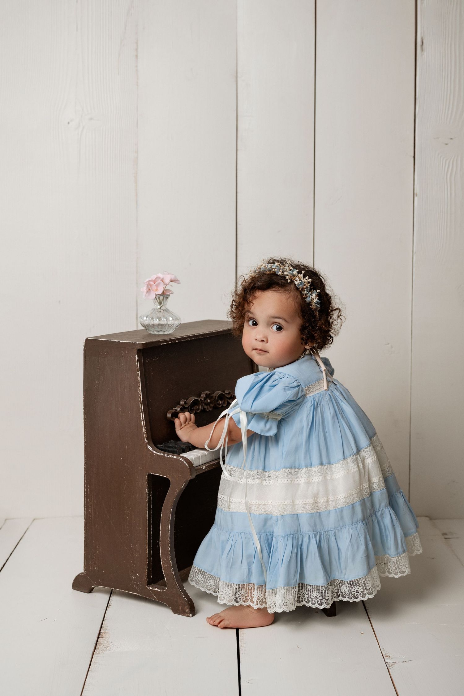 A toddler girl in a blue lace dress with floral crown stands at a vintage toy piano in a white studio.