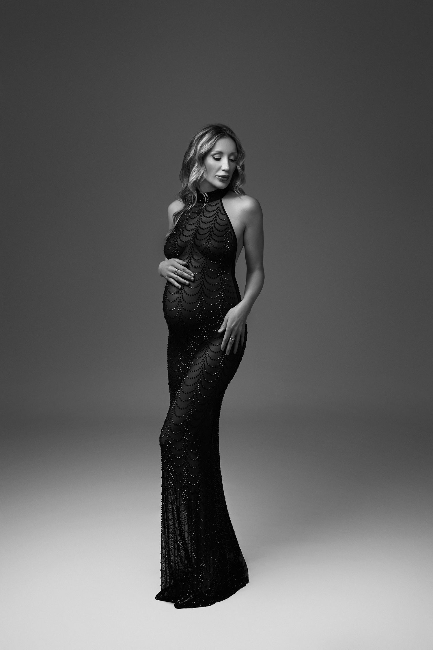 Black and white photo of a pregnant woman in an elegant floor-length sequin gown, posing gracefully in a studio.