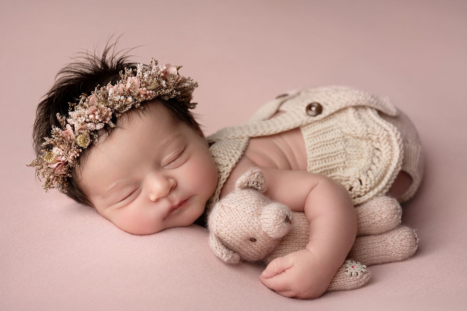 Sleeping newborn baby wearing floral headband cuddles knit teddy bear on pink background.
