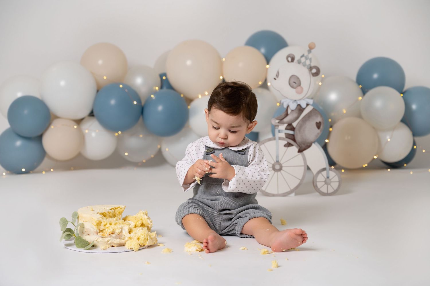 Baby boy in gray romper sits with smash cake surrounded by blue and beige balloons and a robot decoration.