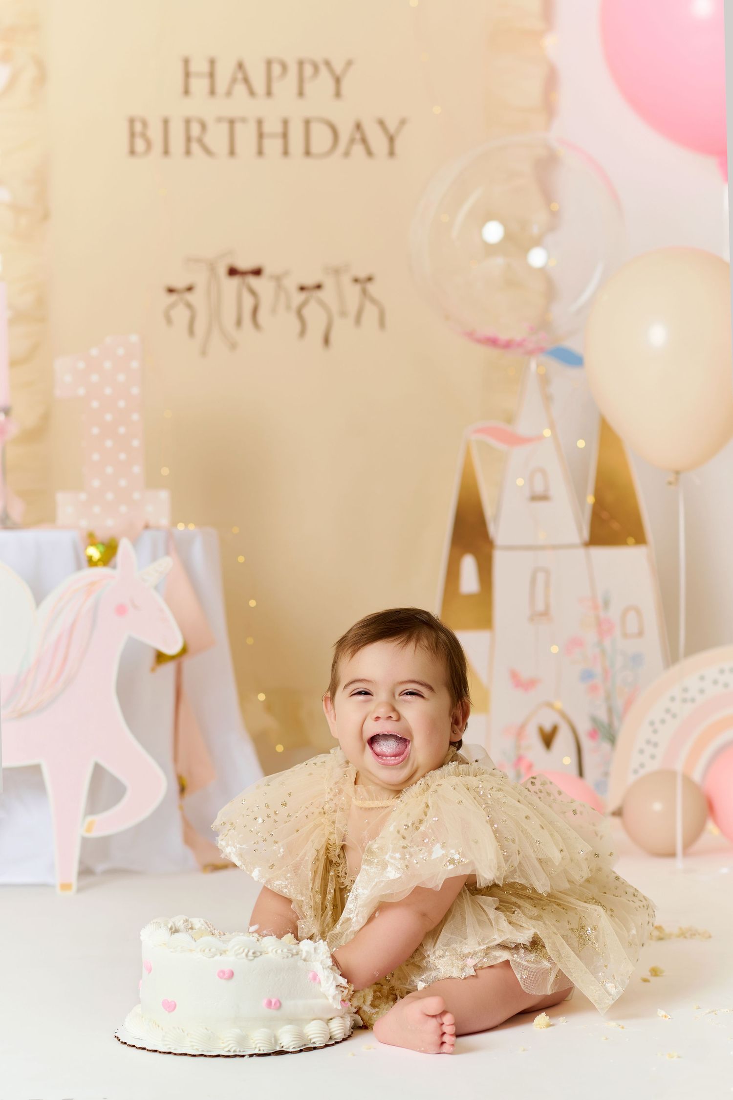 Laughing baby girl in gold dress sits before white smash cake at pink princess birthday party with balloons and decorations.