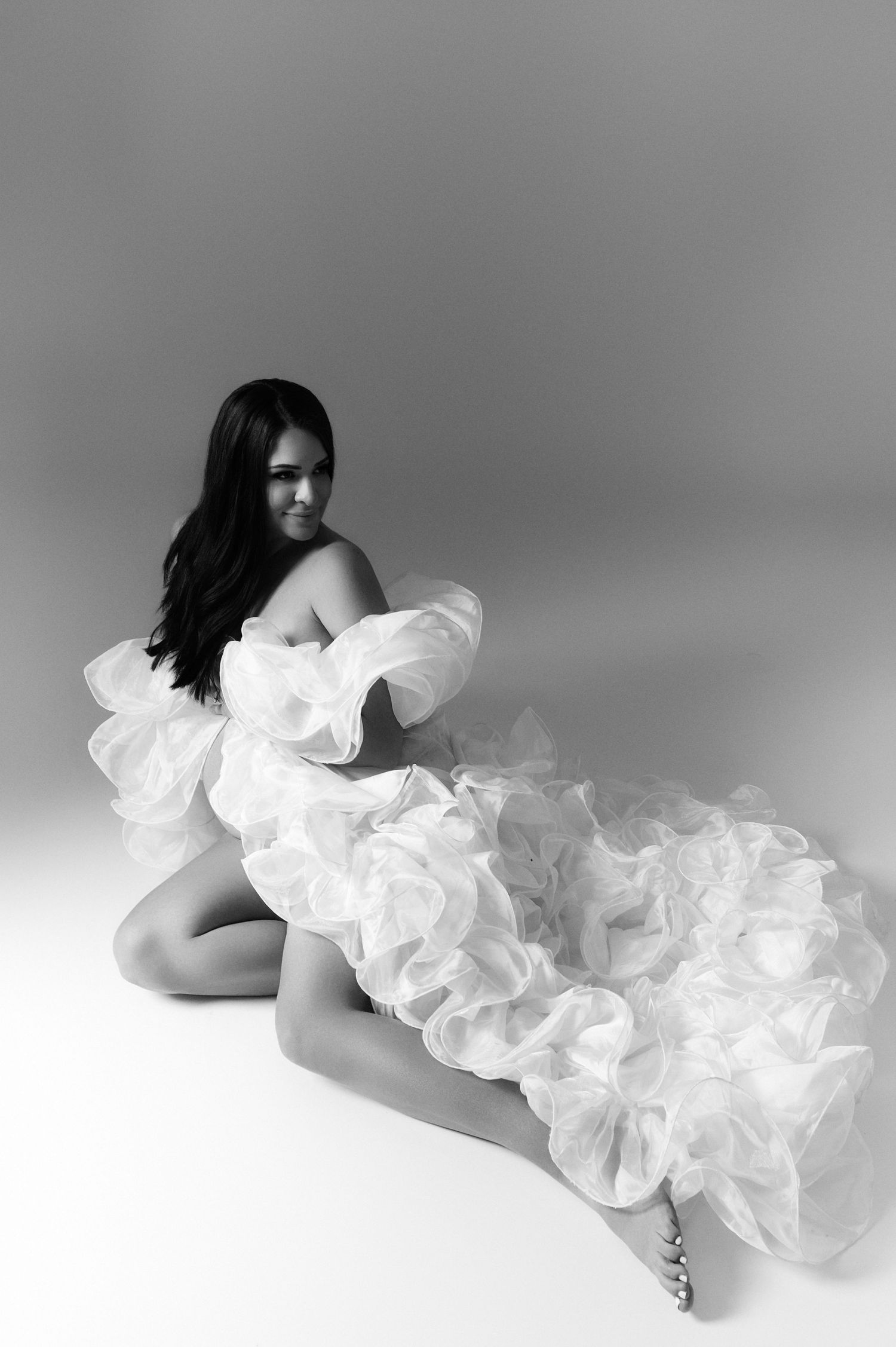 Black and white photo of a woman in a ruffled white gown sitting gracefully on a studio floor.