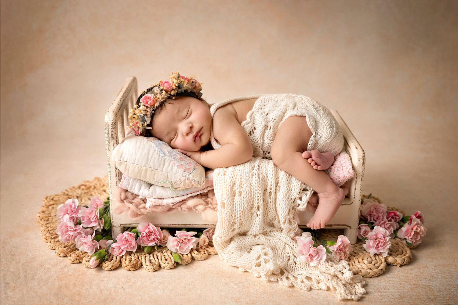 Newborn baby sleeping on a tiny pink bed with floral wreath, lace blanket, and pink roses in a soft studio setting.