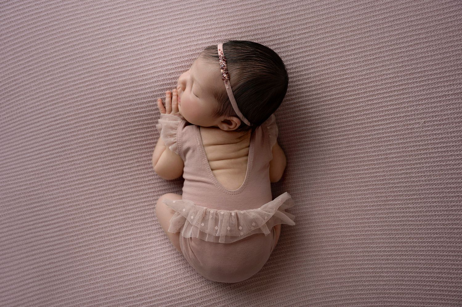 Newborn baby girl sleeping peacefully in pink outfit and headband on soft pink textured backdrop.
